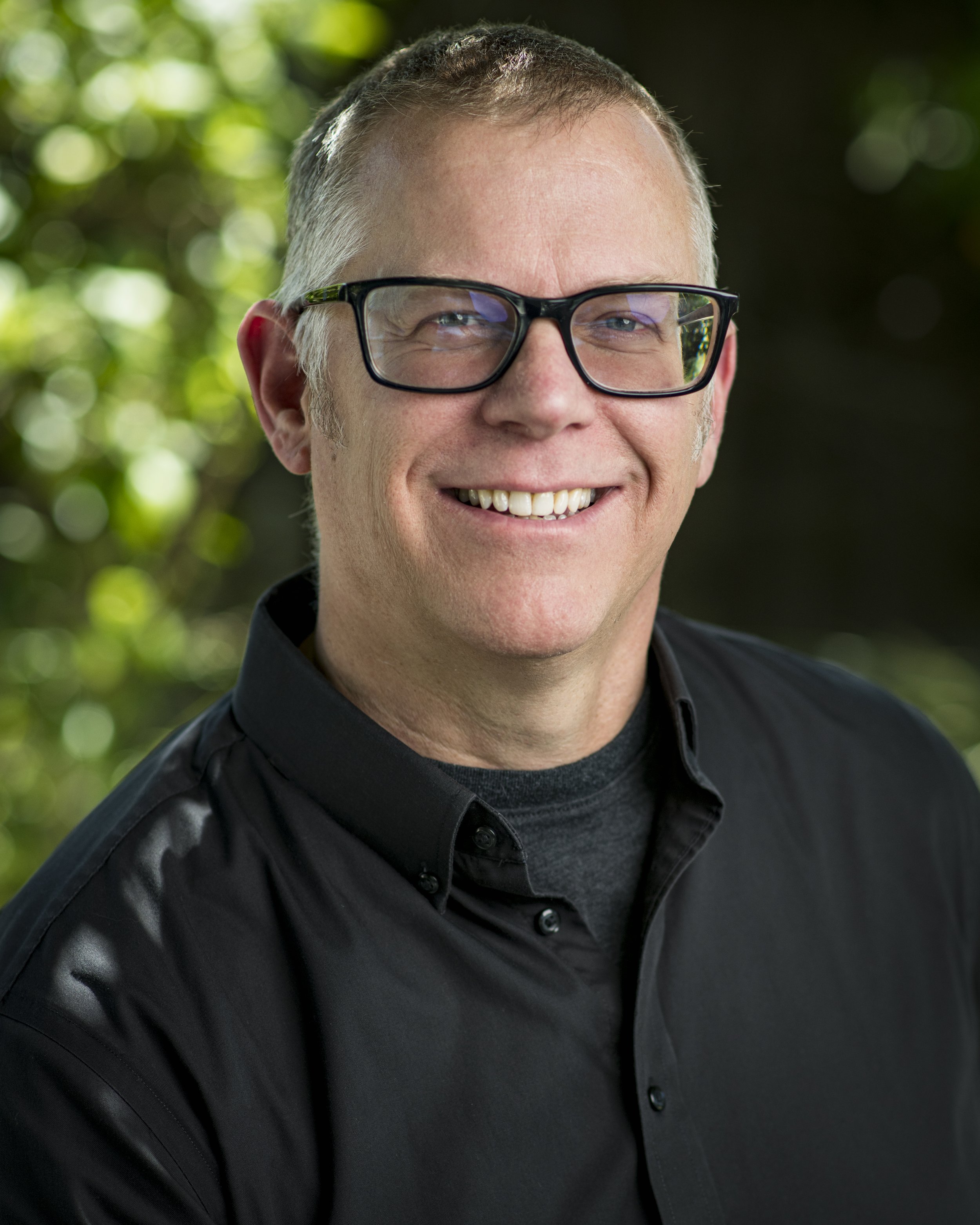 A smiling man with short gray hair, wearing black-rimmed glasses and a black button-up shirt, standing outdoors with a blurred green leafy background.