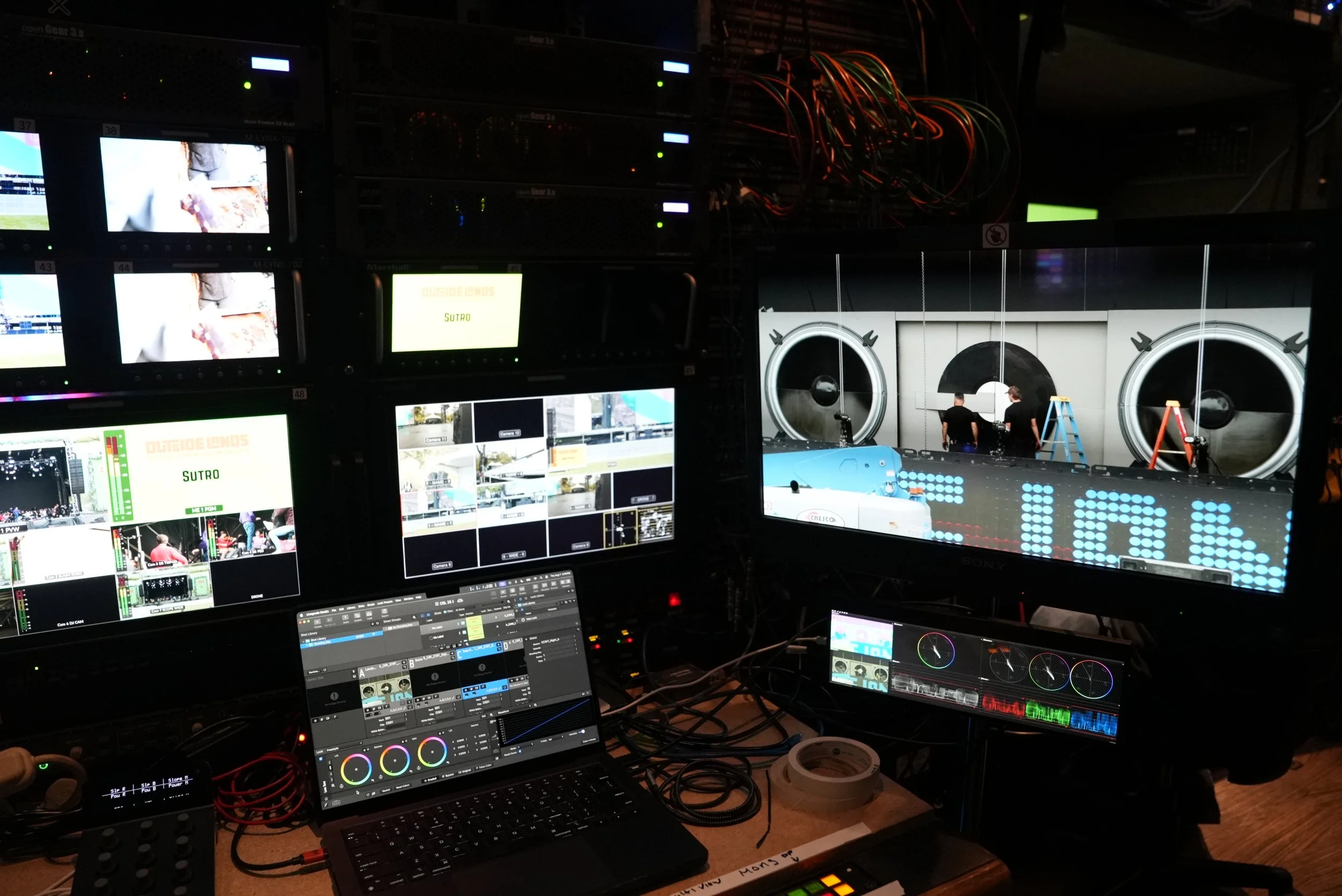 A control room with multiple monitors displaying video feeds, graphics, and monitoring software, likely used for live production or event staging. Behind the screens, a stage is visible with two people setting up equipment in front of large speakers and stage lights.