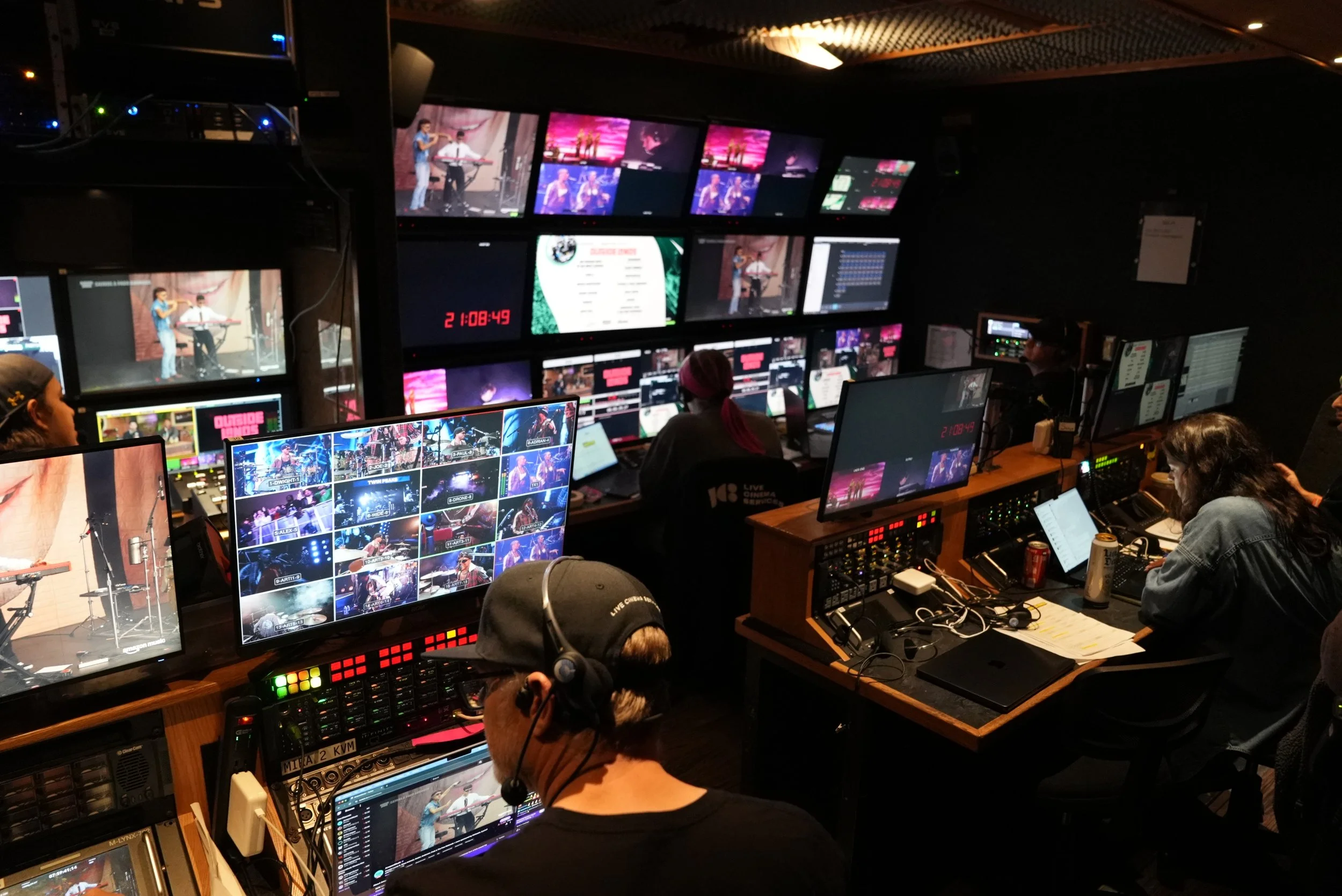 A behind-the-scenes view of a production control room with multiple monitors, operators, and cameras capturing a live event, such as a concert or television broadcast.