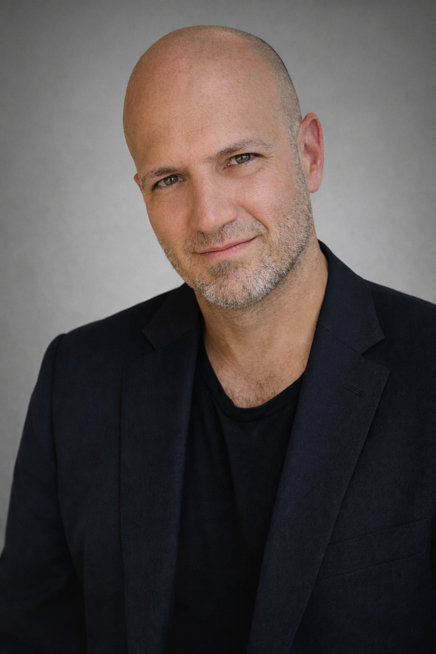 A portrait of a middle-aged, bald man with a short beard, wearing a black shirt and blazer, looking directly at the camera with a slight smile against a neutral gray background.