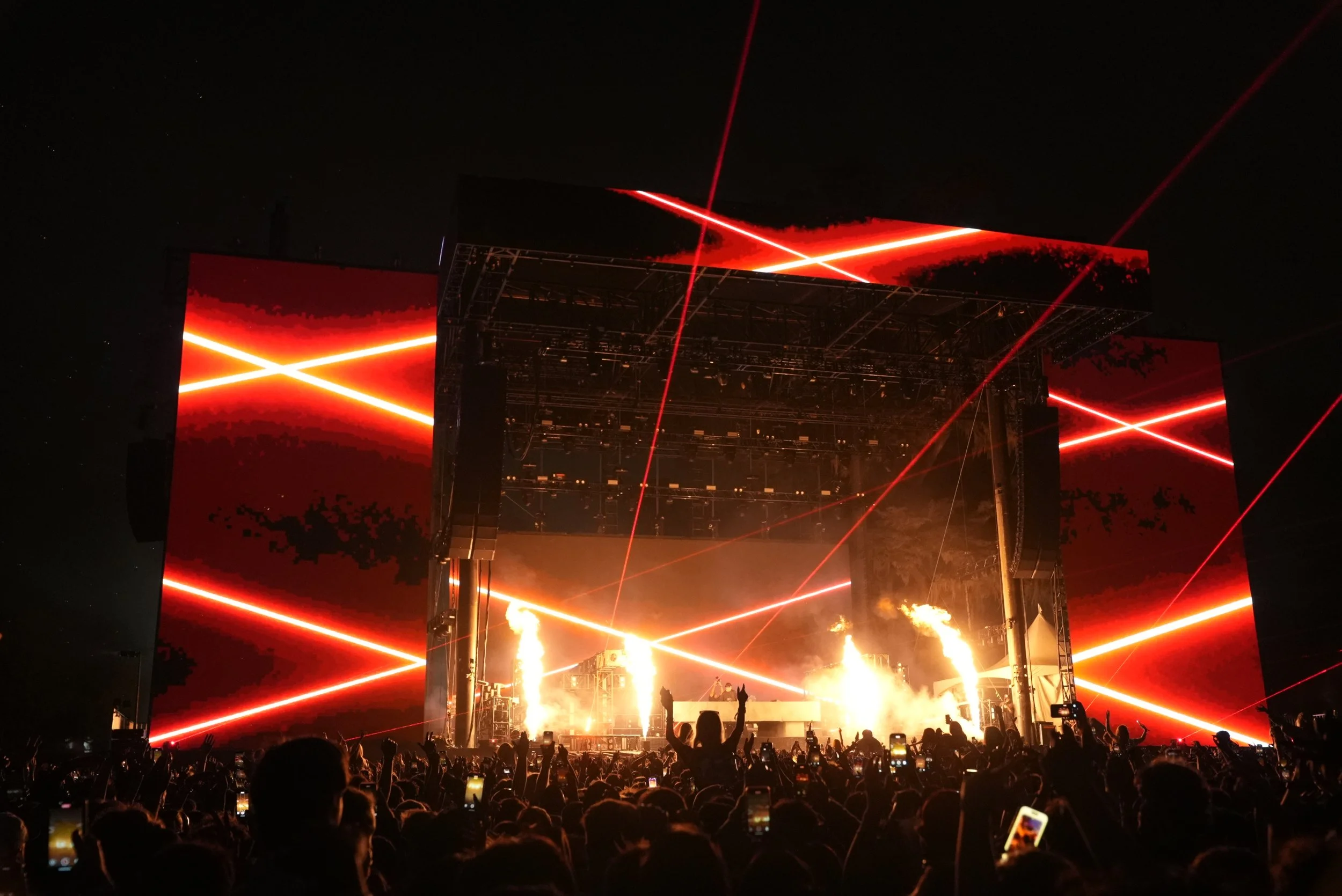 Nighttime concert scene with large stage, laser lights, fire effects, and a crowd of people holding up phones.