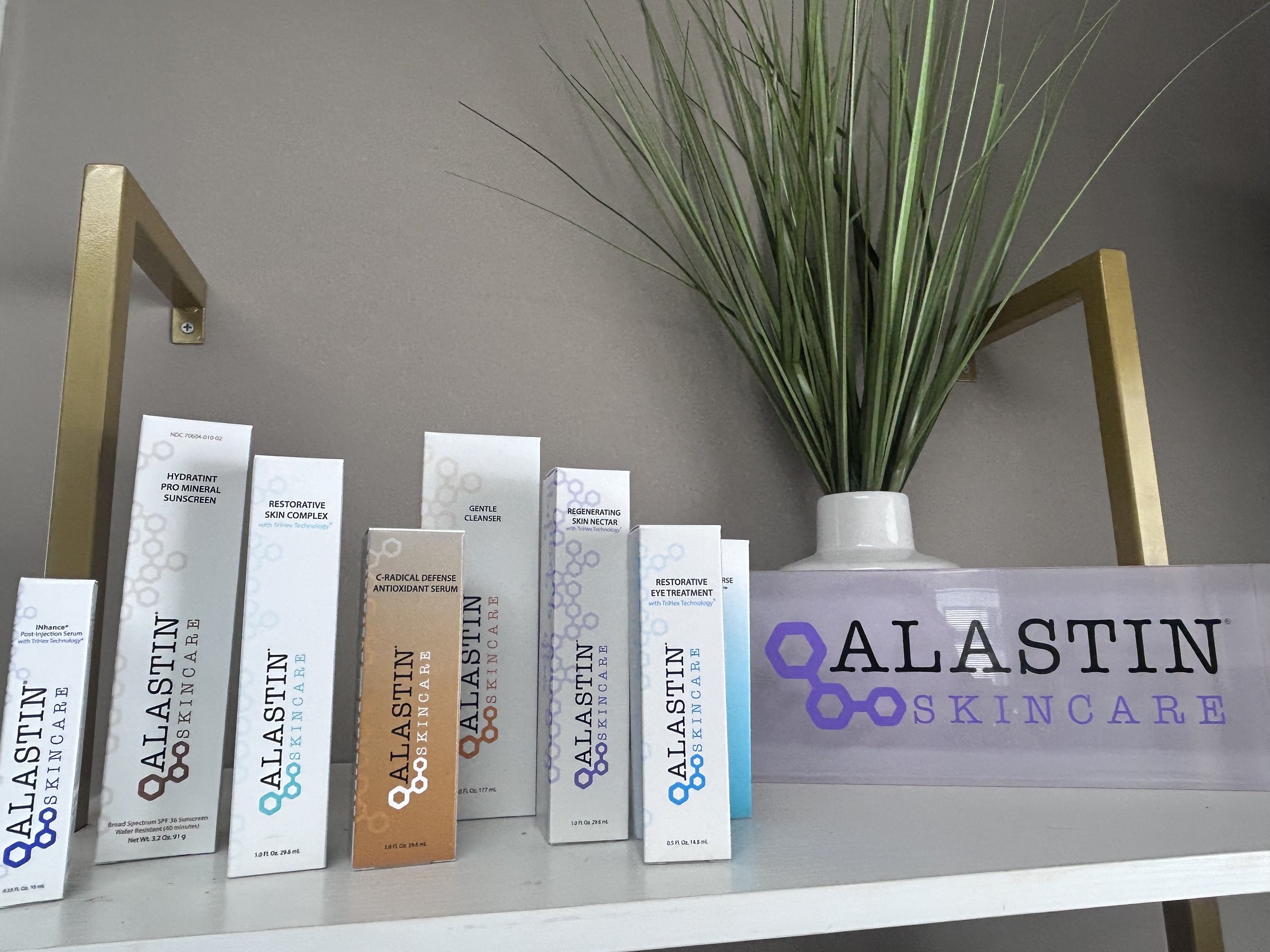 Skincare products from Alastin on a white shelf, with a decorative green plant in a white vase and a wooden sign with the Alastin logo in purple.