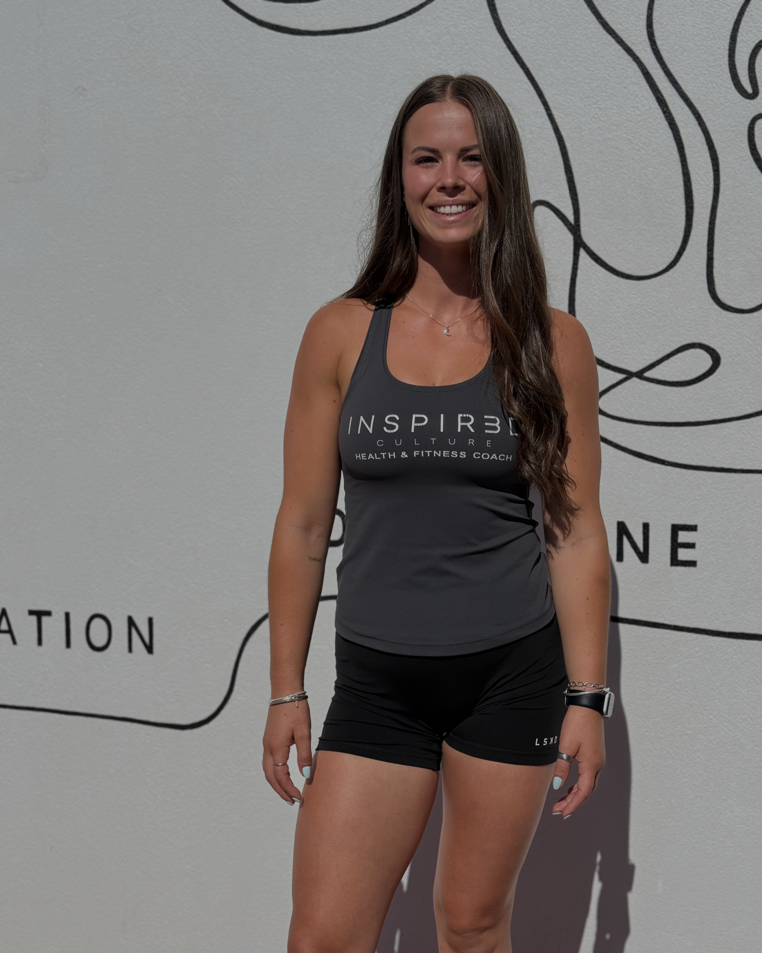 A young woman with long brown hair wearing a black tank top and black shorts, smiling and standing in front of a wall with a black abstract line drawing and text.