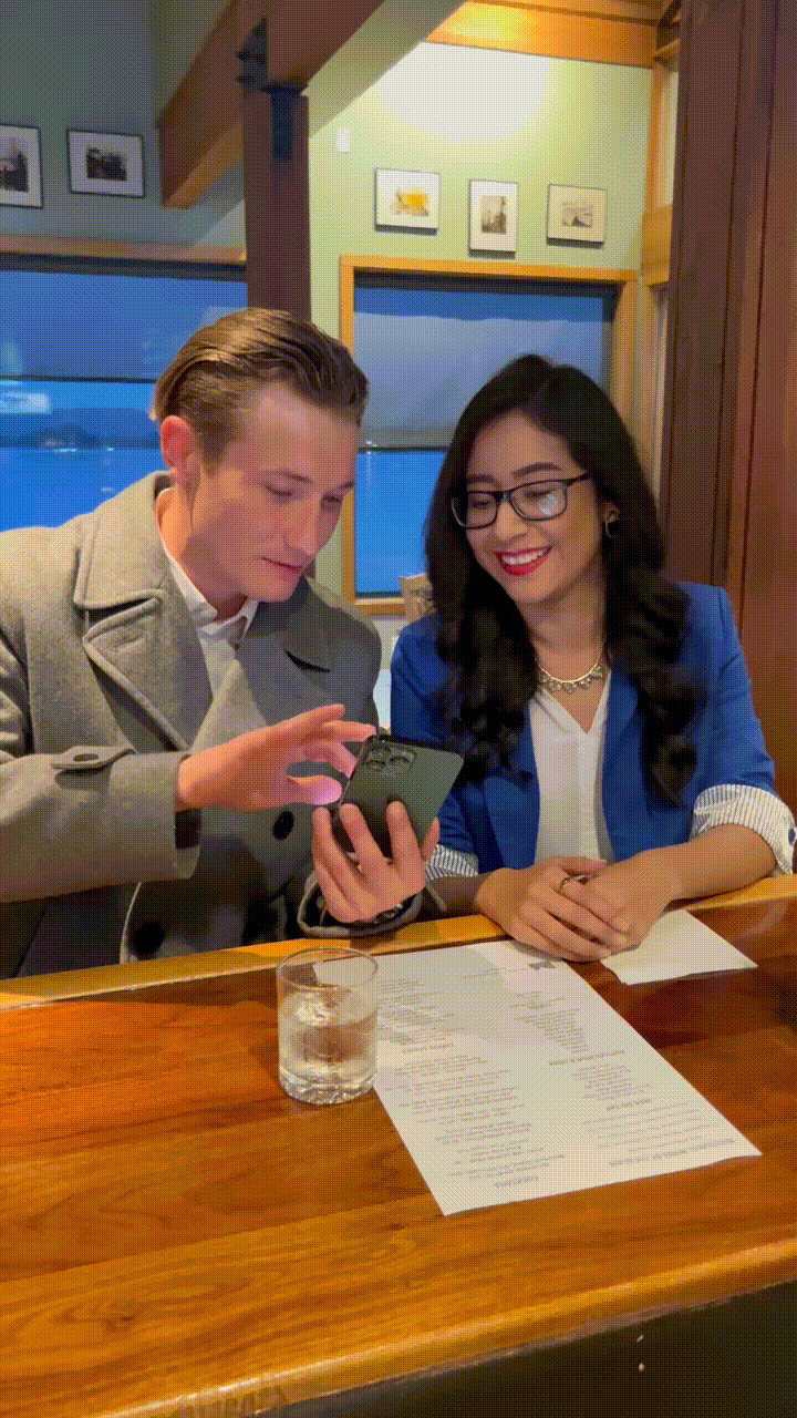 A man and woman sitting at a wooden table in a cozy restaurant, looking at a phone together. There are papers and a glass of water on the table.