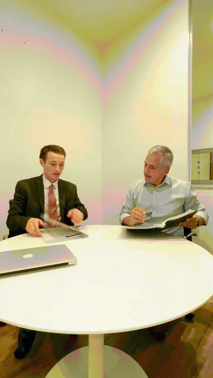 Two men sitting at a white oval table having a discussion in a small office. One man, young and in a suit, is holding a magazine or folder, while the older man is holding papers and a pen.
