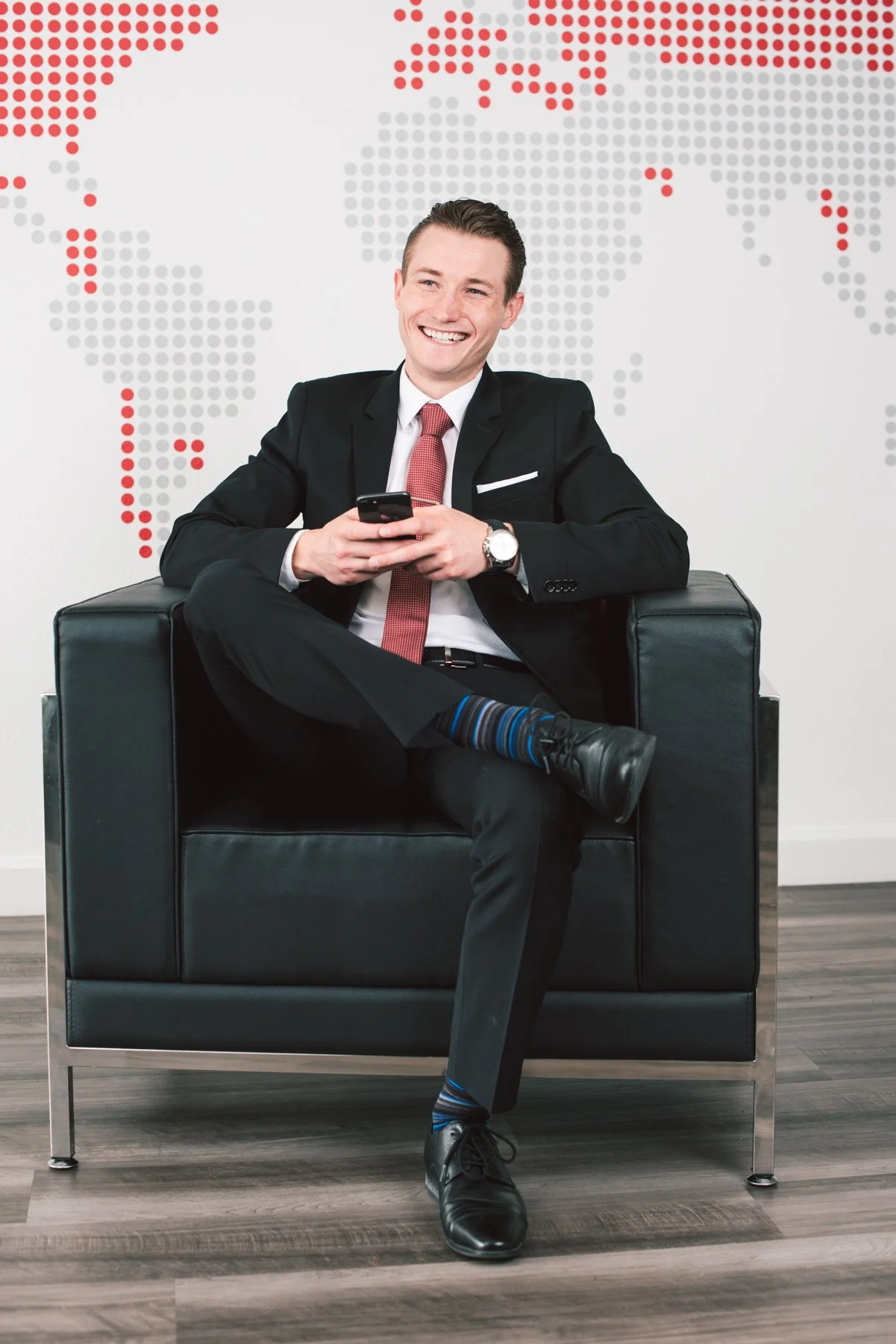 A man in a business suit sitting on a black armchair, smiling and looking at his phone, with a world map made of dots on the white wall behind him.