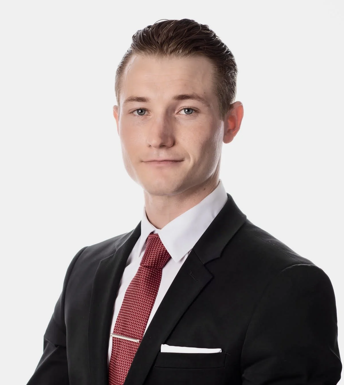 A professional young man with light skin, styled brown hair, wearing a black suit, white shirt, and red tie, smiling slightly at the camera against a plain white background.