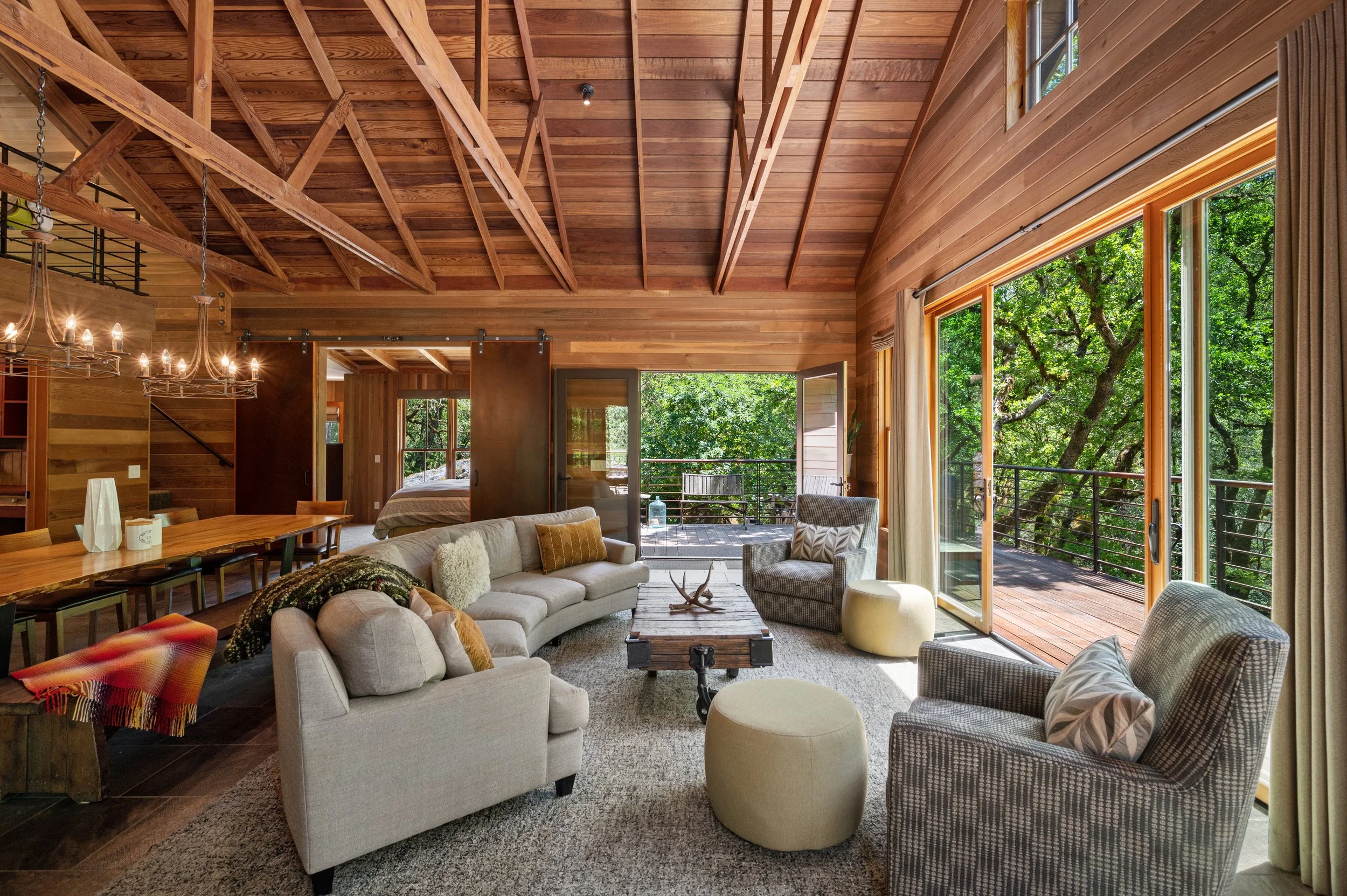 Interior of a spacious living room with wooden walls, high vaulted ceiling with exposed beams, large windows, and a sliding glass door leading to a balcony with trees outside. The room has a beige sofa, patterned chairs, a coffee table, and a chandelier.