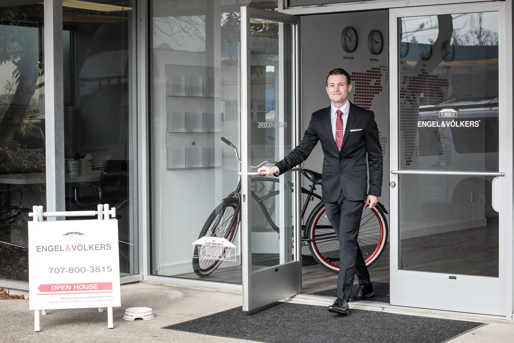 Man in a suit holding the door open at a real estate office with bicycles inside, advertising for Engel & Völkers, with an open house sign outside.