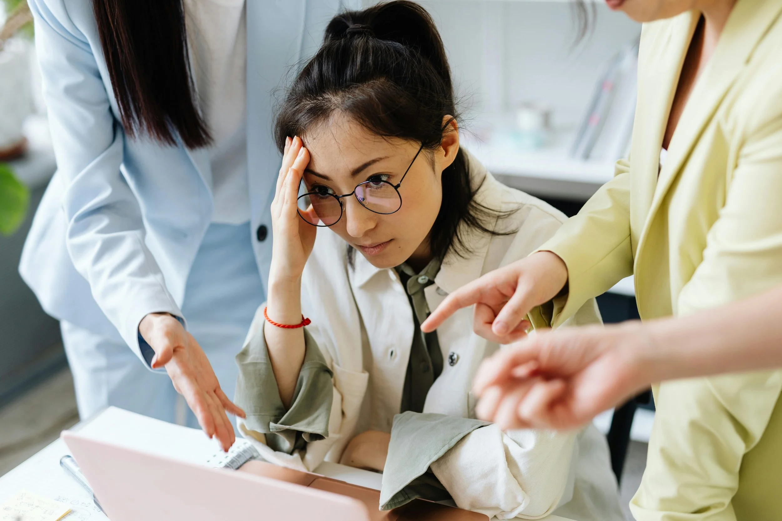 Professional woman sitting at a desk looking overwhelmed while colleagues point at a laptop, representing constant demands and a state of response in the workplace