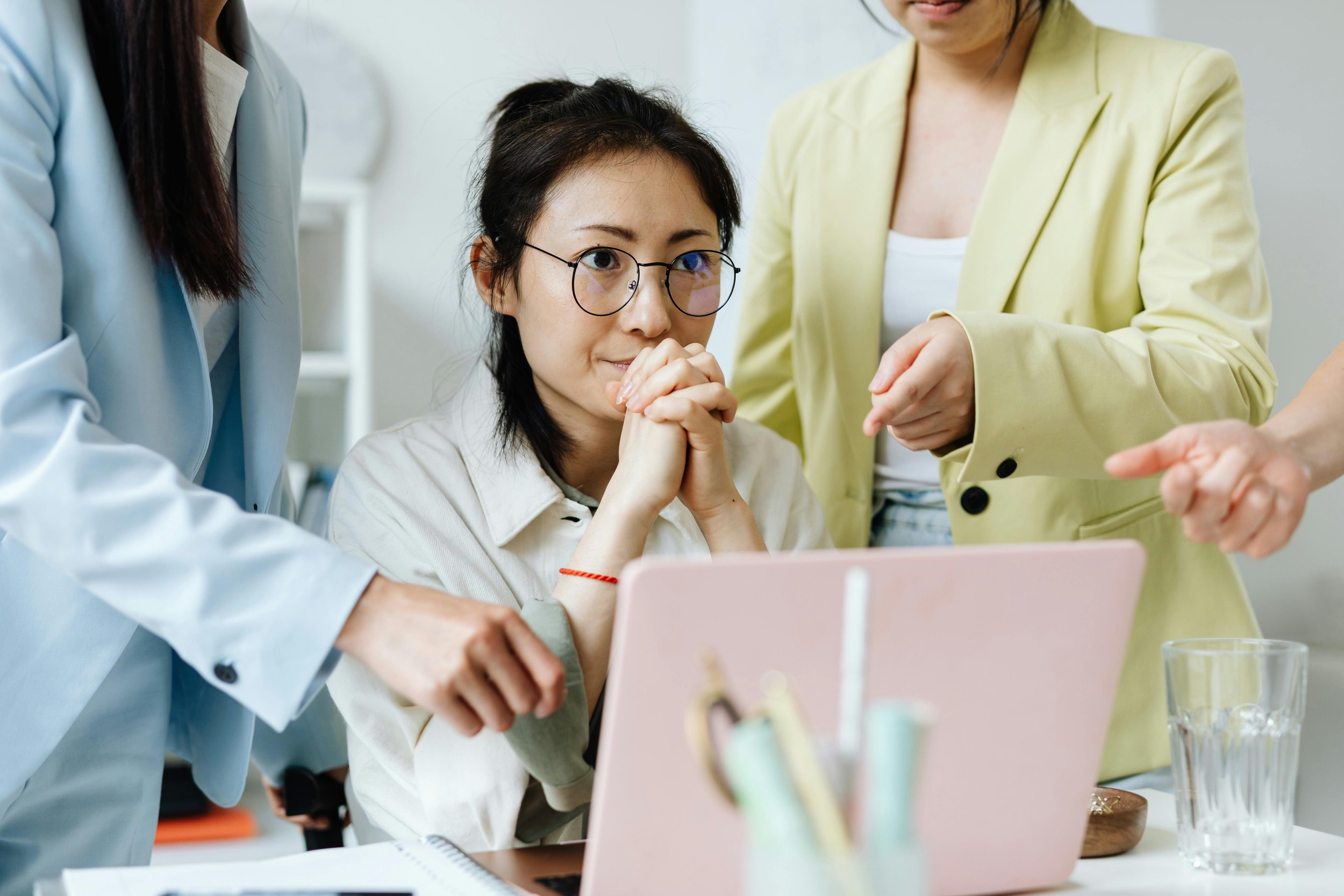 Professional woman sitting at a desk with hands clasped, appearing stressed while others gesture around her, representing pressure, decision fatigue, and workplace overwhelm