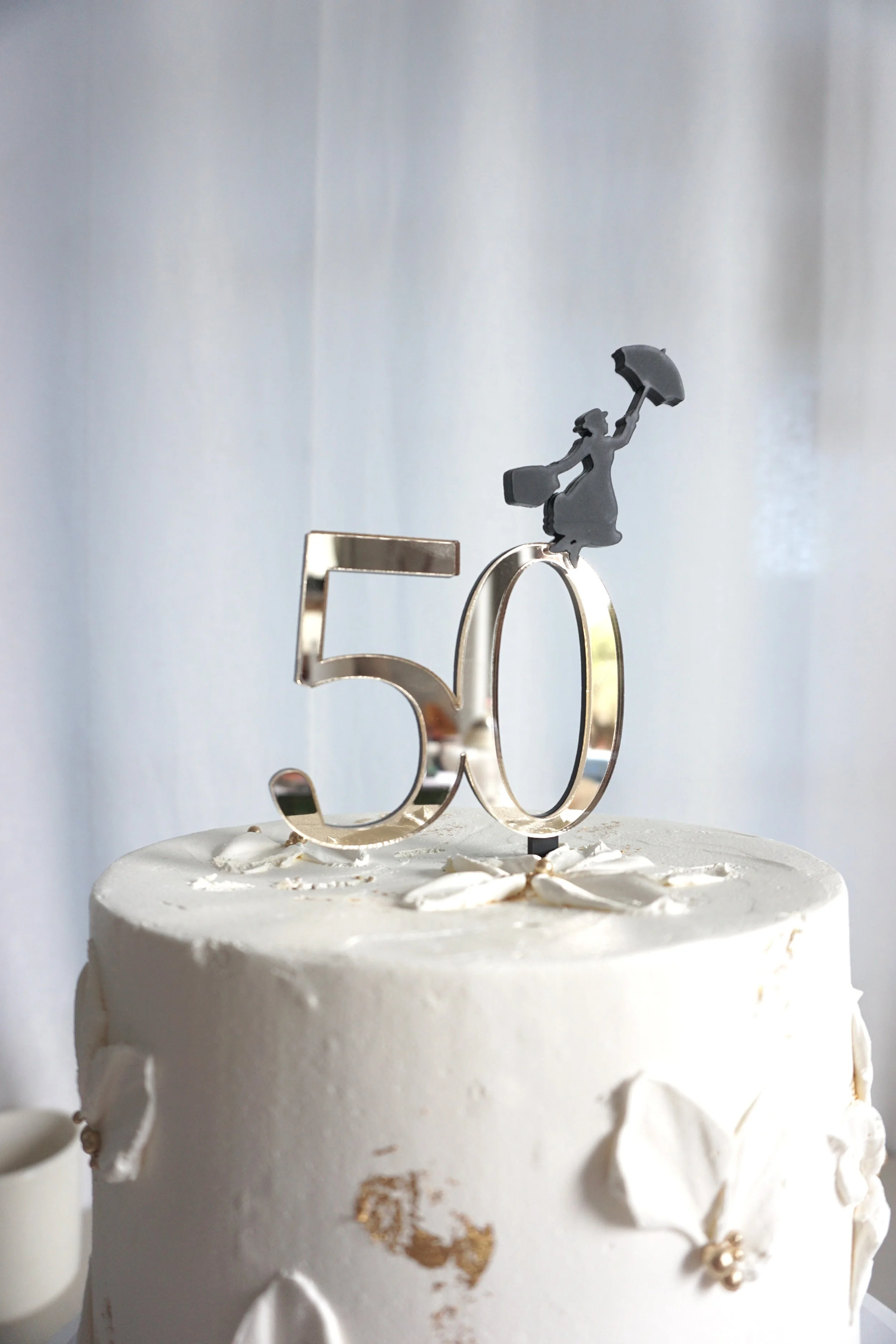 A cake topper celebrating a 50th birthday and uses Mary Poppins as a theme. Features Mary Poppins as a silhouette about to fly off the gold mirror 50 cake topper