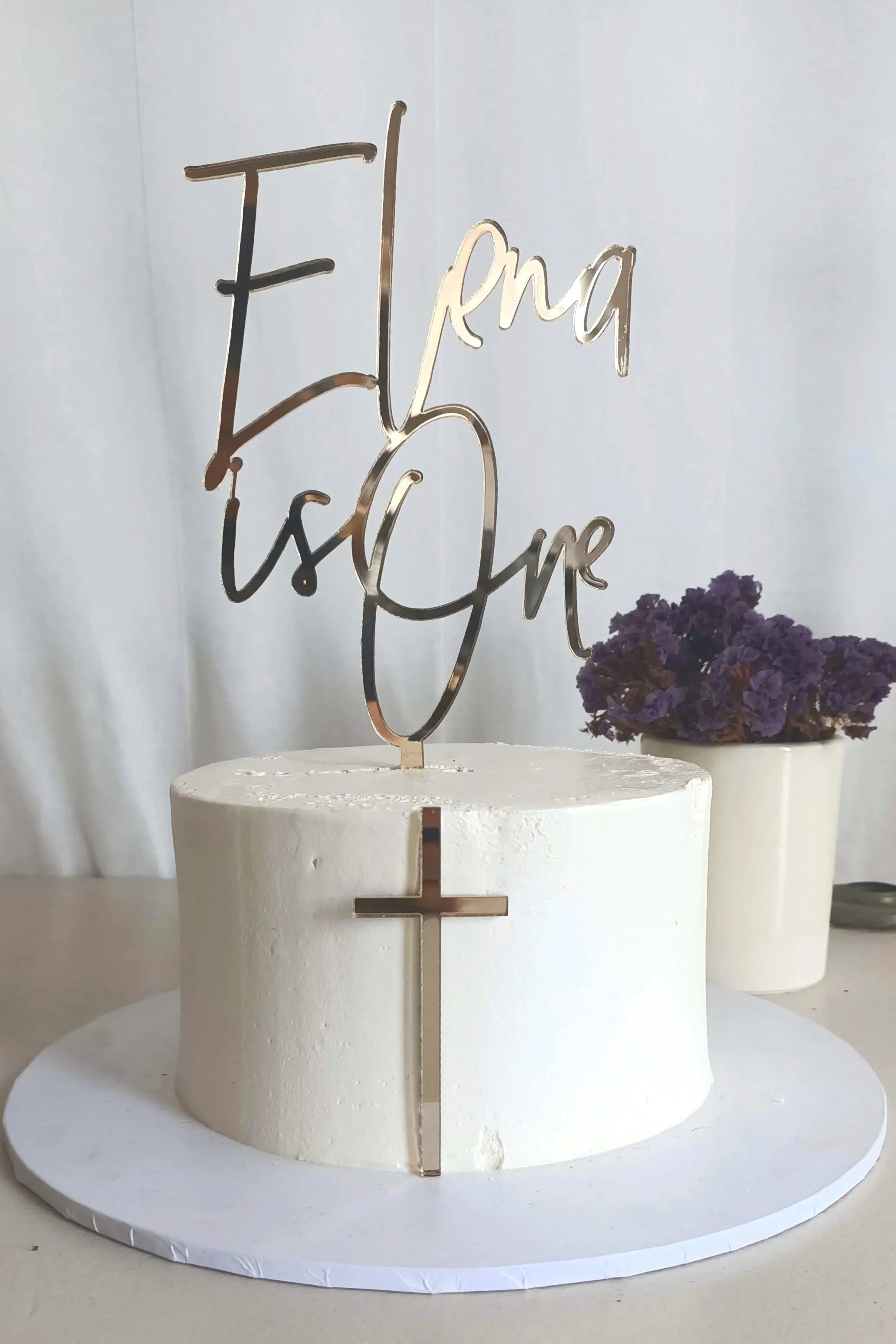A small golden cross used as a fropper in combination with the Delilah Style cake topper which reads Elena is One. This can be used for birthdays, baptisms, and other religious events