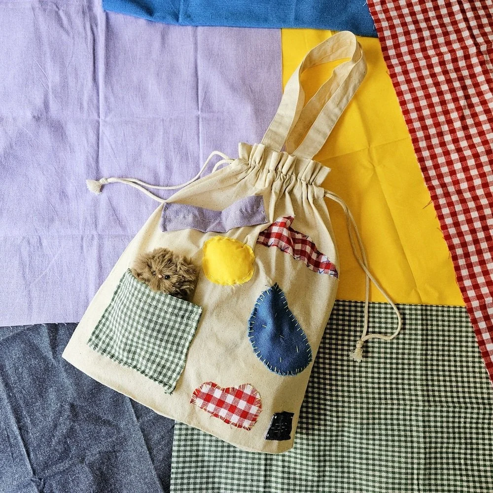 Fabric tote bag with a small fluffy animal peeking out of a square pocket, surrounded by colorful fabric pieces in shades of purple, yellow, red checkered, green checkered, and black checkered.