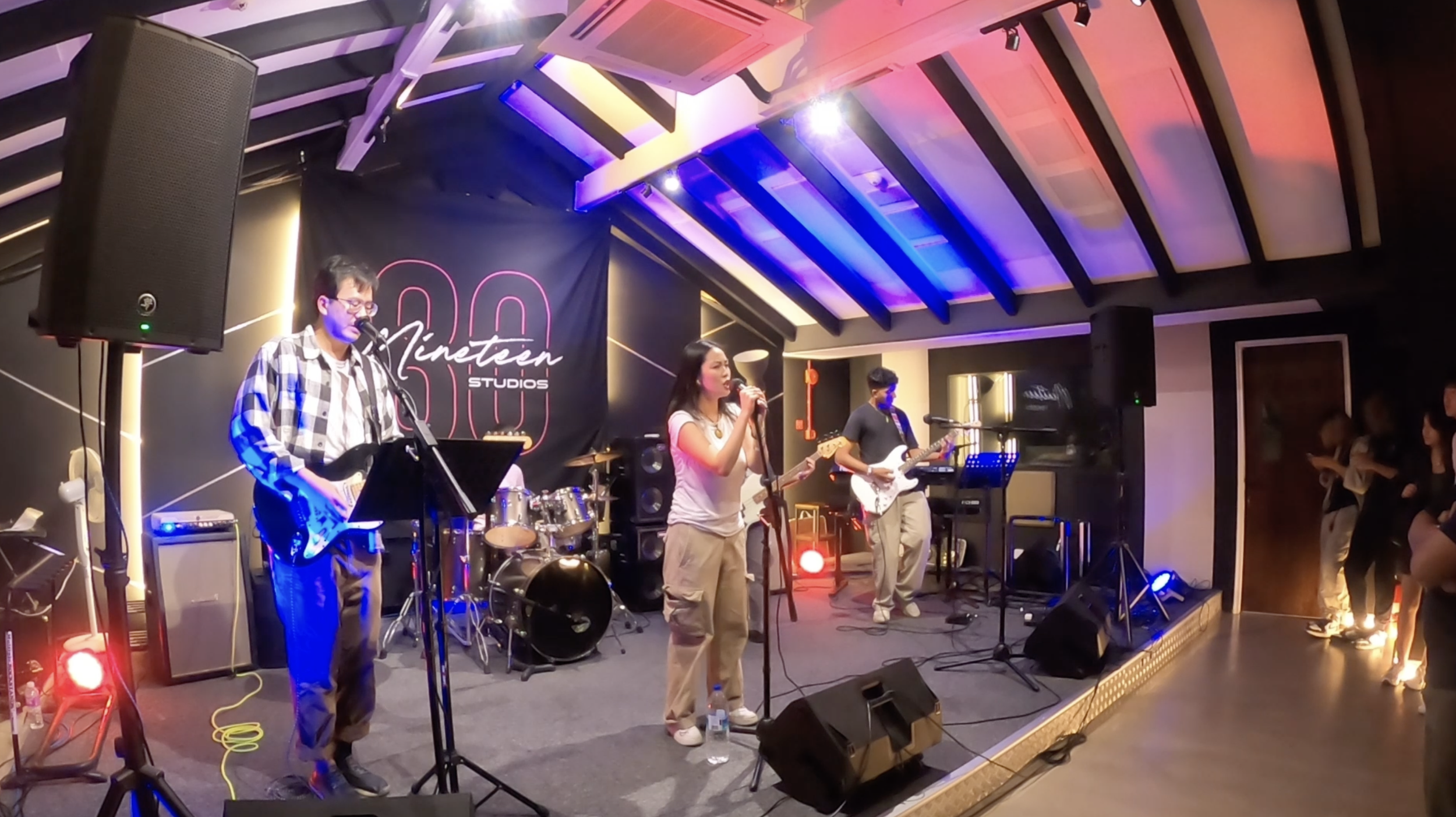 A band performing live on stage at a music studio, with a singer, guitarists, a drummer, and audience members in the background.