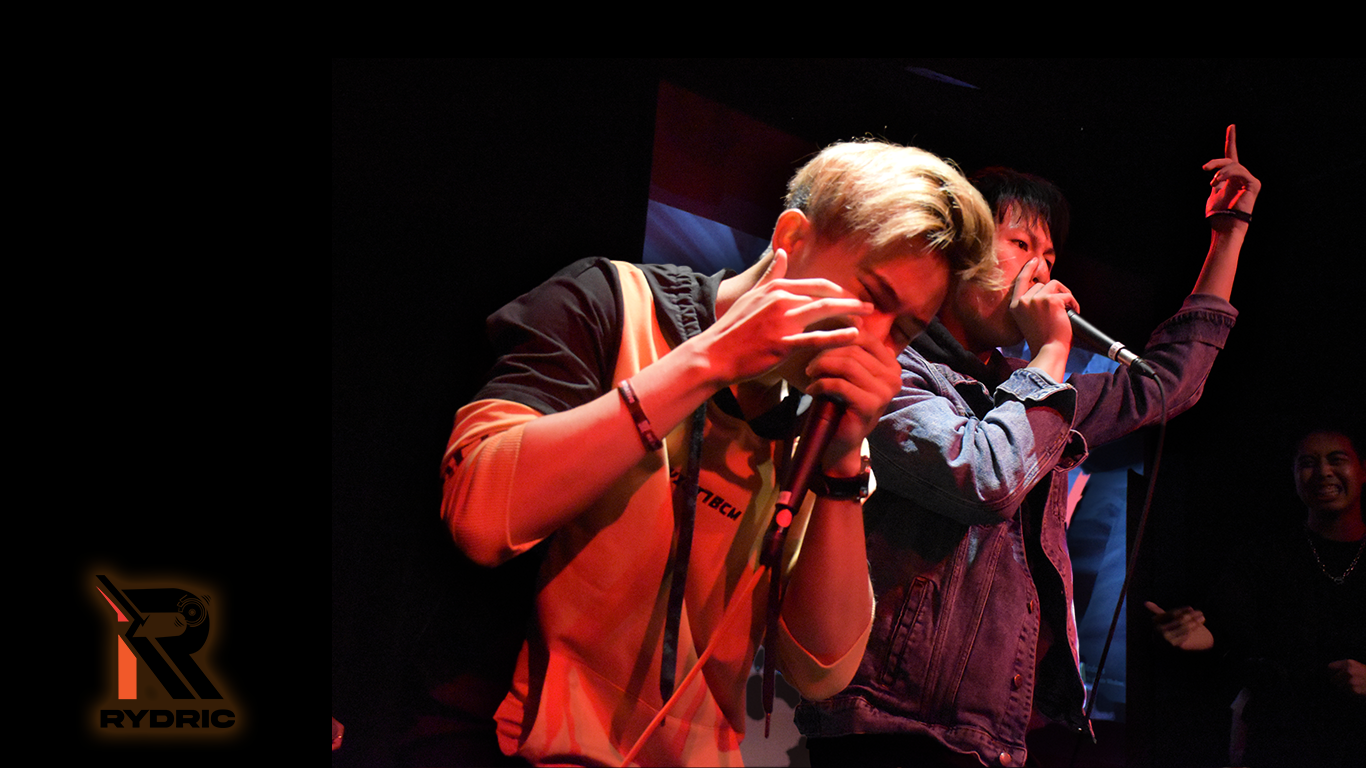 Two young men performing on stage with microphones, one with blonde hair and the other with dark hair, both passionately singing or shouting, with a woman smiling in the background.