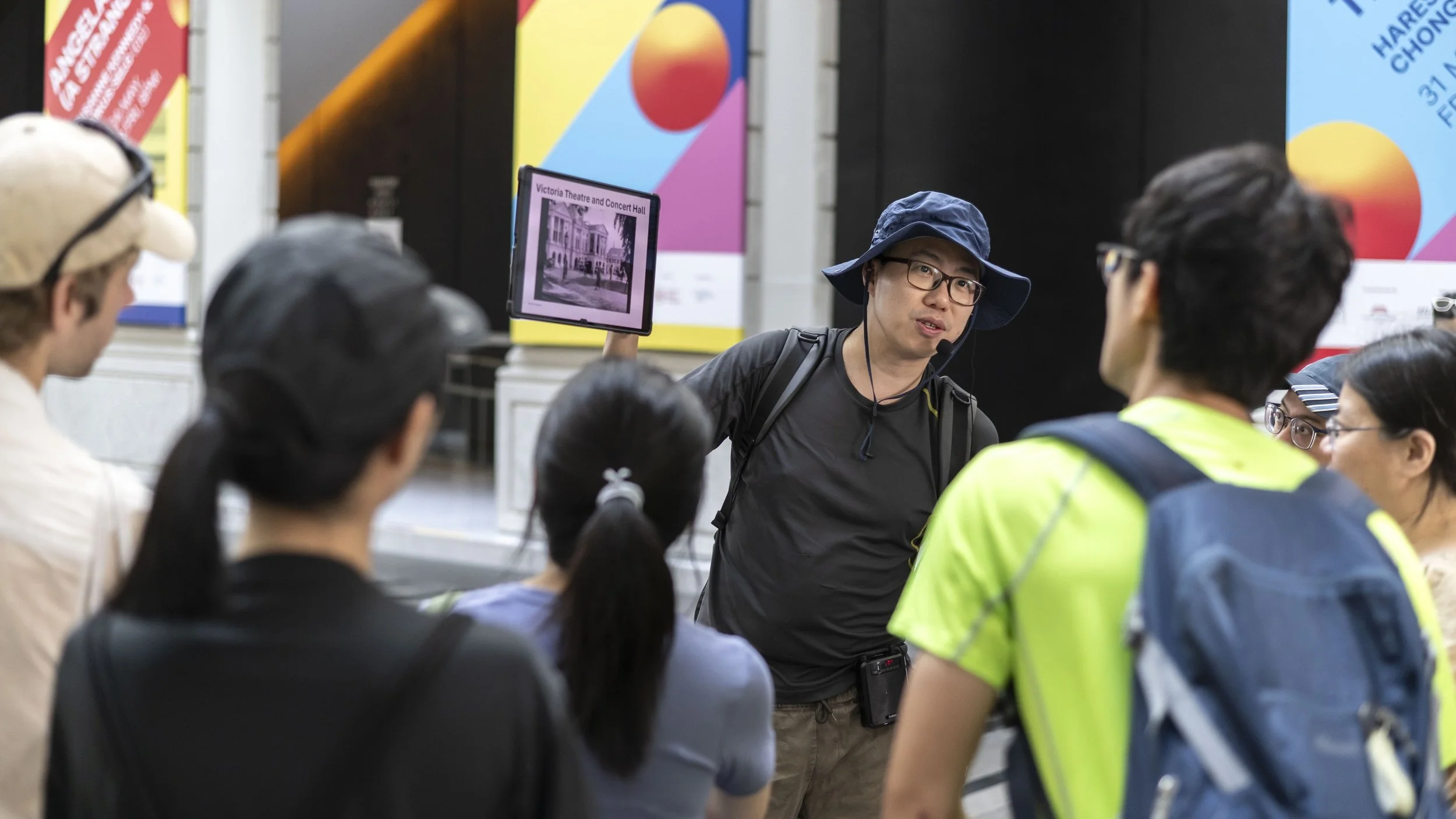 A tour guide in a blue hat and glasses giving a talk to a group of diverse tourists at a museum, with colorful posters in the background.