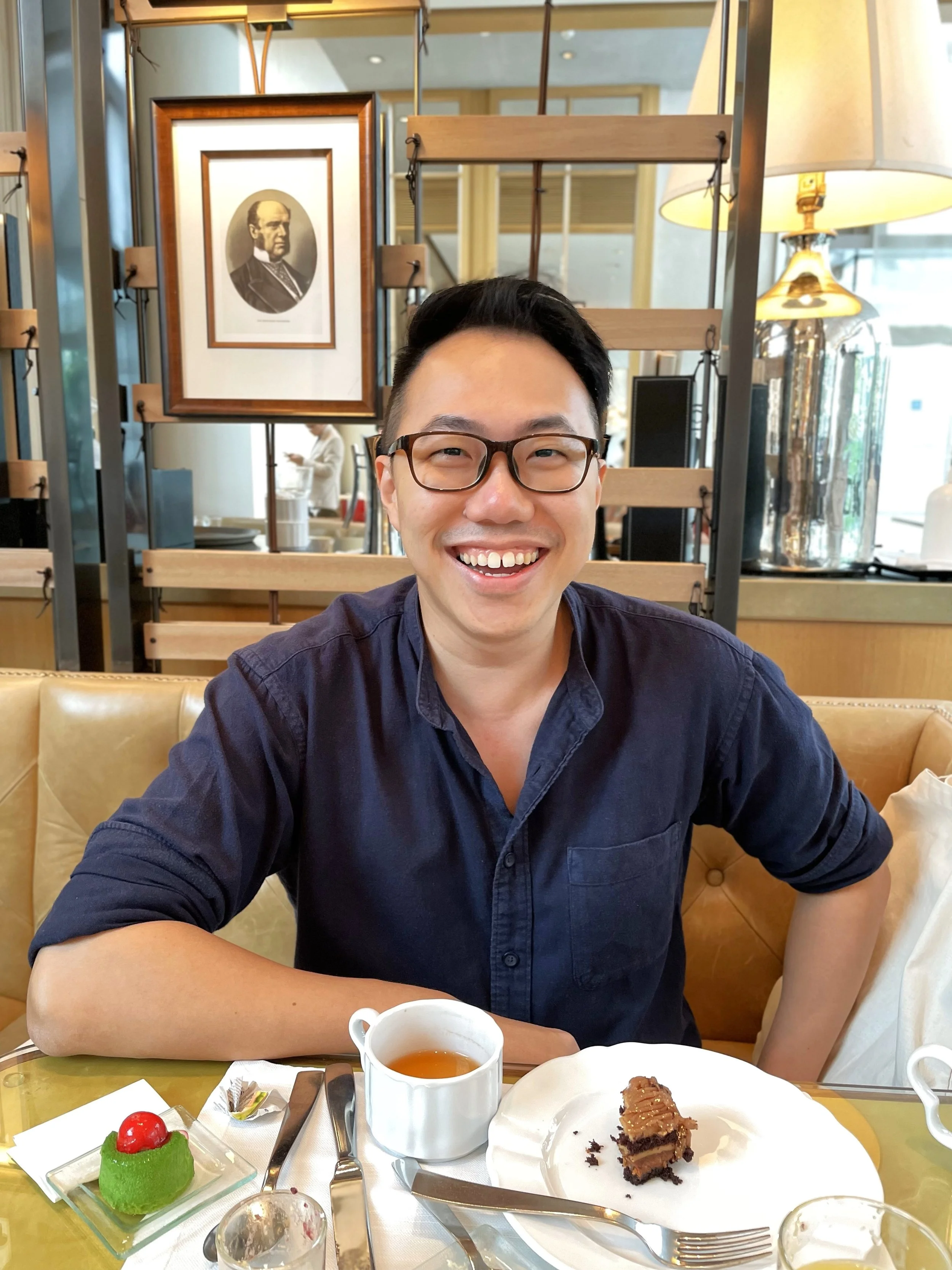 A man with black hair and glasses smiling at a table in a restaurant or cafe, with a slice of chocolate cake, a cup of tea, and a green dessert topped with a cherry in front of him.