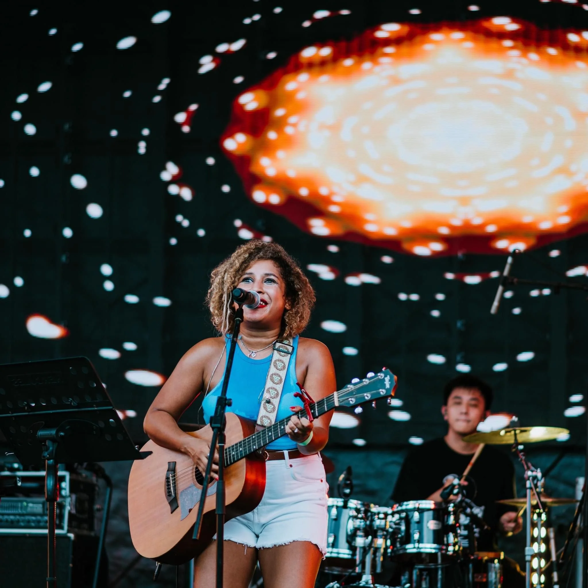 A woman with curly hair singing into a microphone while playing an acoustic guitar on stage, with a drummer in the background and a colorful, abstract digital display overhead.