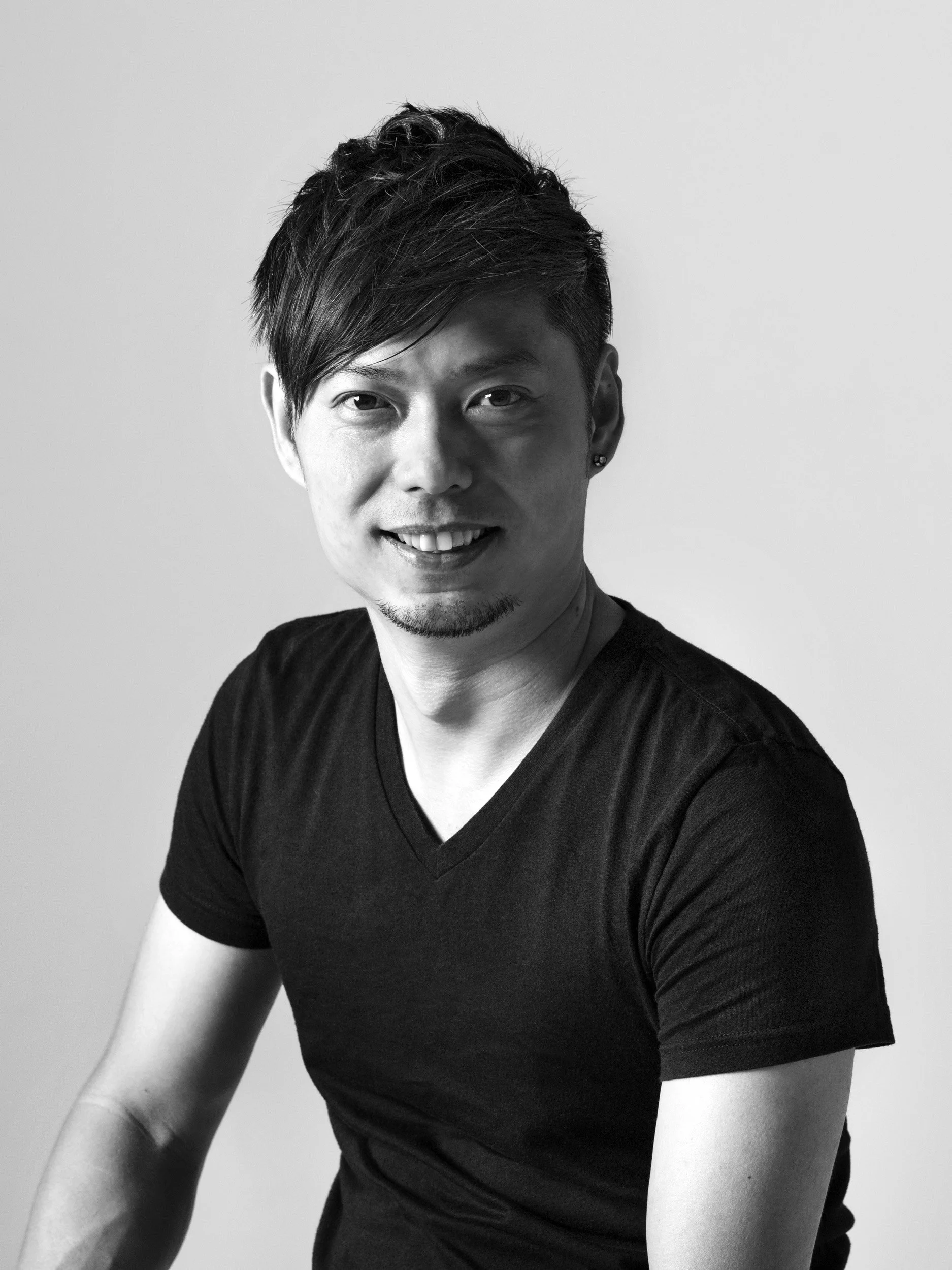 Black and white portrait of a young man with styled hair, wearing a black V-neck T-shirt, smiling at the camera against a plain background.