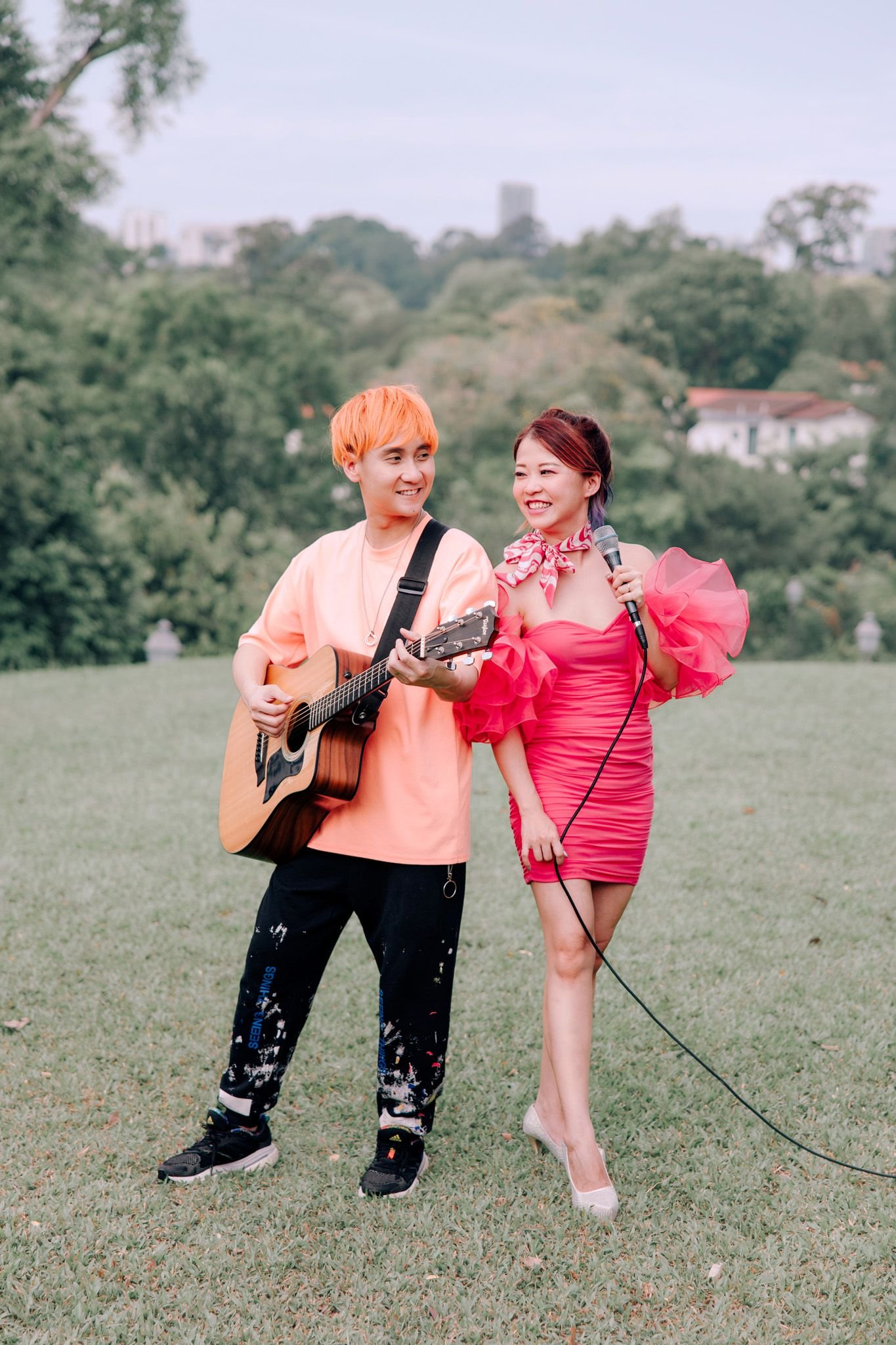 Two women performing outdoors, one playing guitar and the other singing into a microphone, surrounded by green trees and a cloudy sky.