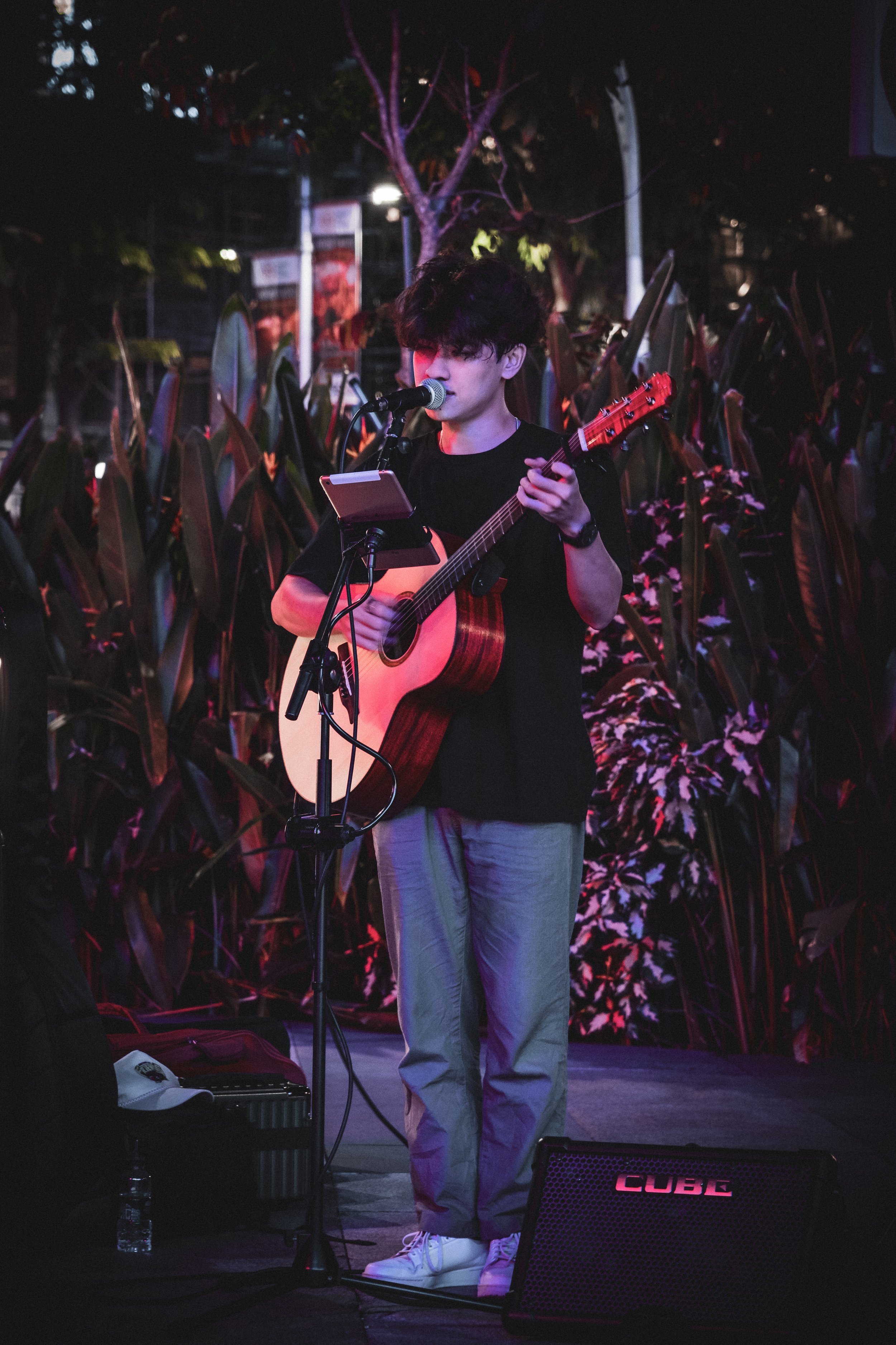 A young man performing on stage at night, playing an acoustic guitar and singing into a microphone, with tall plants in the background.