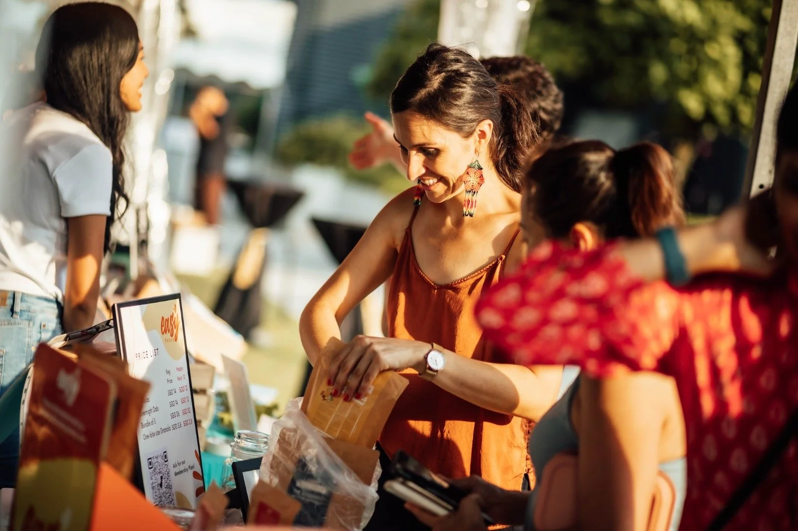 A woman in an orange sleeveless top and colorful earrings is smiling as she shops at an outdoor market stall, engaging with a vendor. Other people are visible in the background, and the scene takes place during daylight.