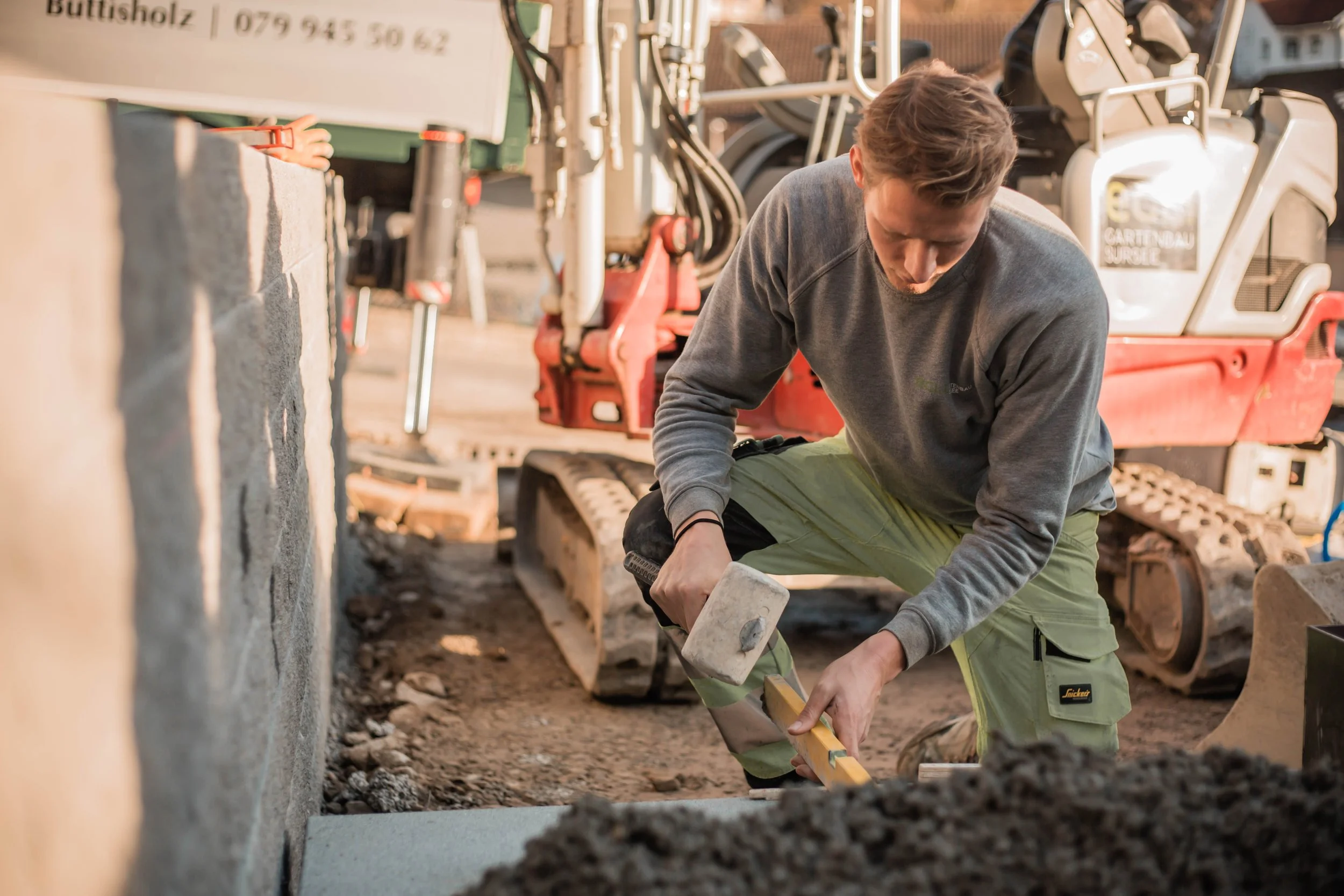 Mann arbeitet auf einer Baustelle, benutzt einen Hammer und ein Meißel, während im Hintergrund ein Minibagger steht.