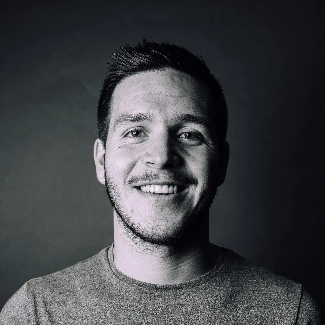 Schwarz-weiße Aufnahme eines lachenden jungen Mannes mit kurzen dunklen Haaren und Bart, trägt ein graues T-Shirt.
