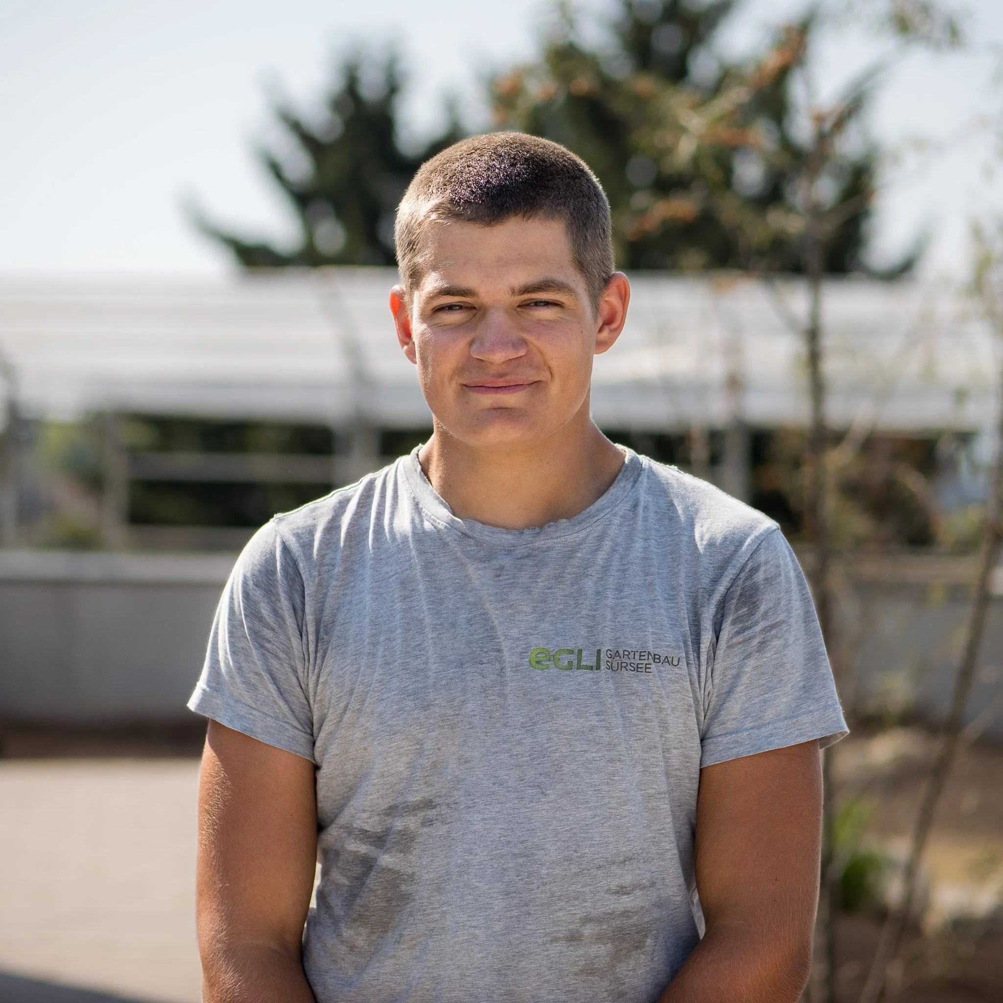 Junge männliche Person steht im Freien während des Tages, trägt ein graues T-Shirt mit grüner und schwarzer Aufschrift, im Hintergrund sind Bäume und Gebäudestrukturen sichtbar.