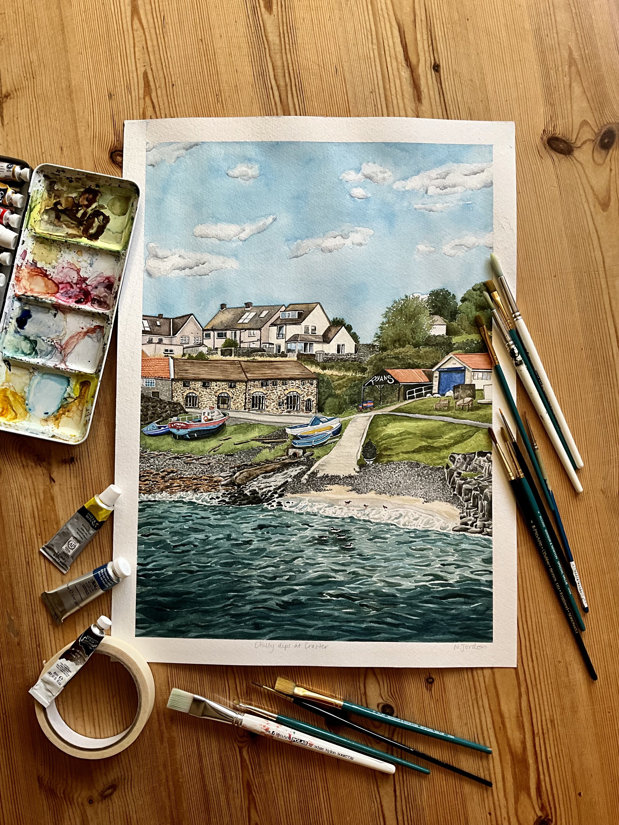 Original A3 ‘Chilly dips at Craster’ watercolour painting (portrait orientation), laid out on a wooden table and surrounded by art materials