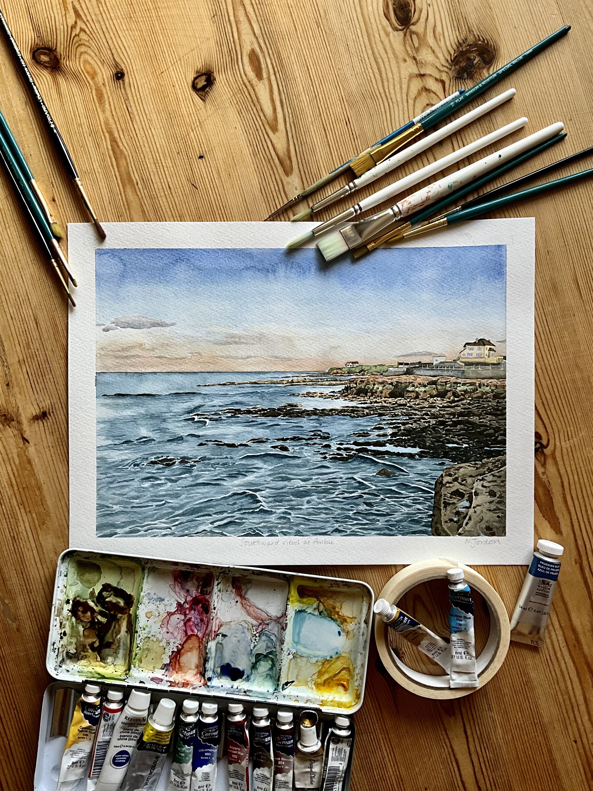 Original A4 ‘Southward views from Amble’ watercolour painting, laid on wooden table and surrounded by watercolour painting supplies