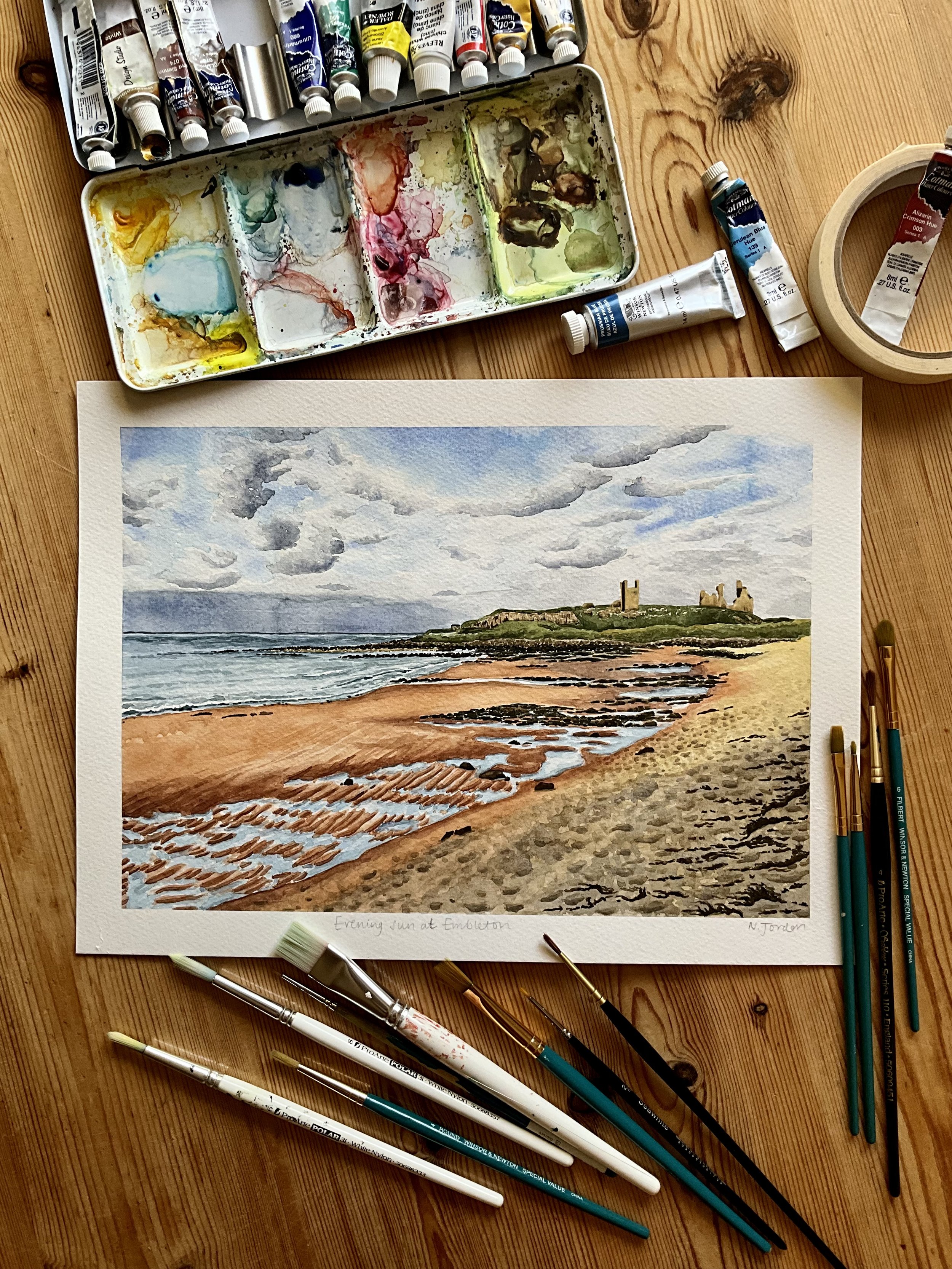 Original A4 ‘Evening sun at Embleton’ watercolour painting. Laid out on wooden table and painting is surrounded by watercolour supplies.