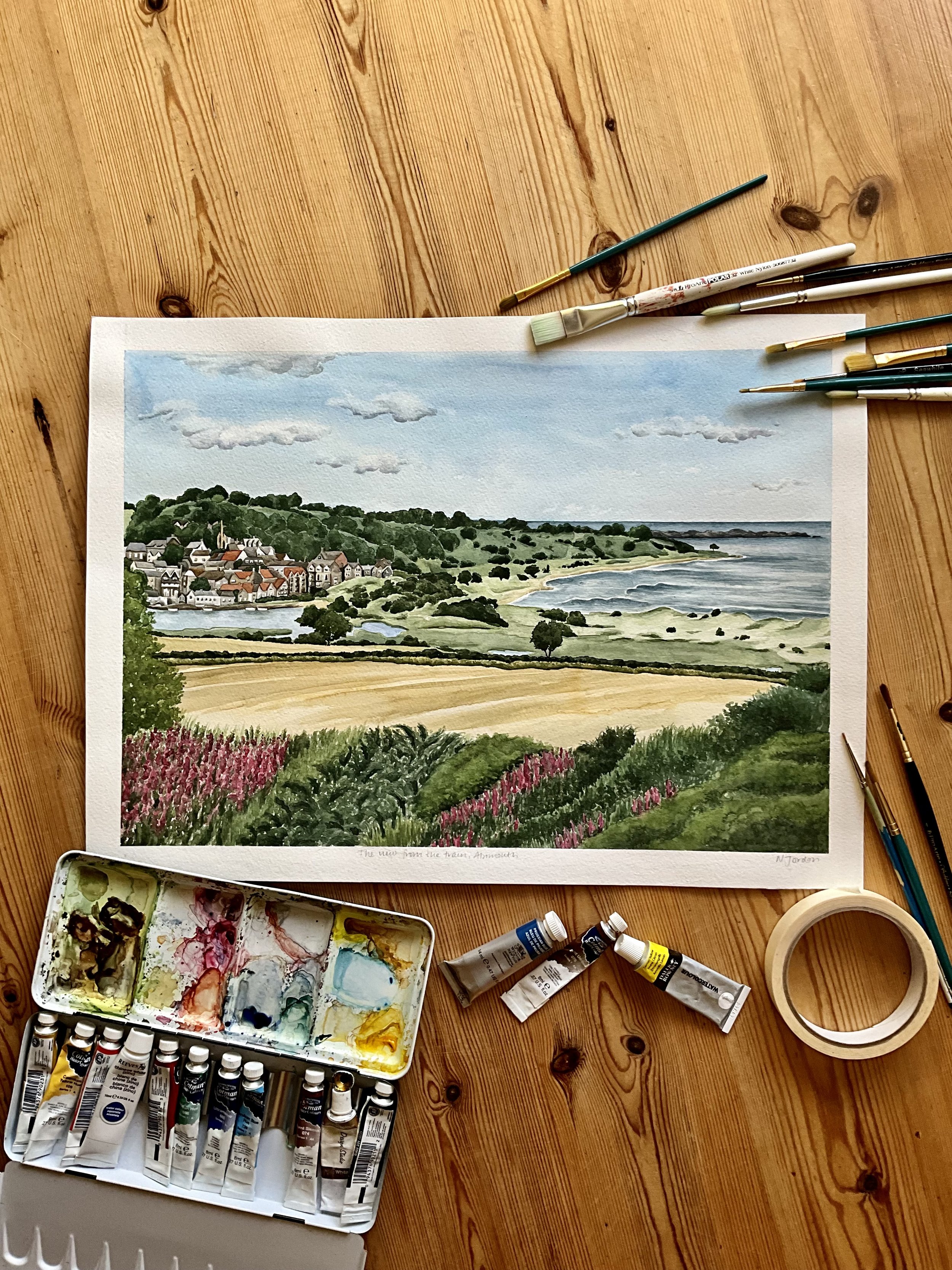 Original A3 ‘The view from the train, Alnmouth’ watercolour painting. Laid on wooden table and surrounded by watercolour supplies