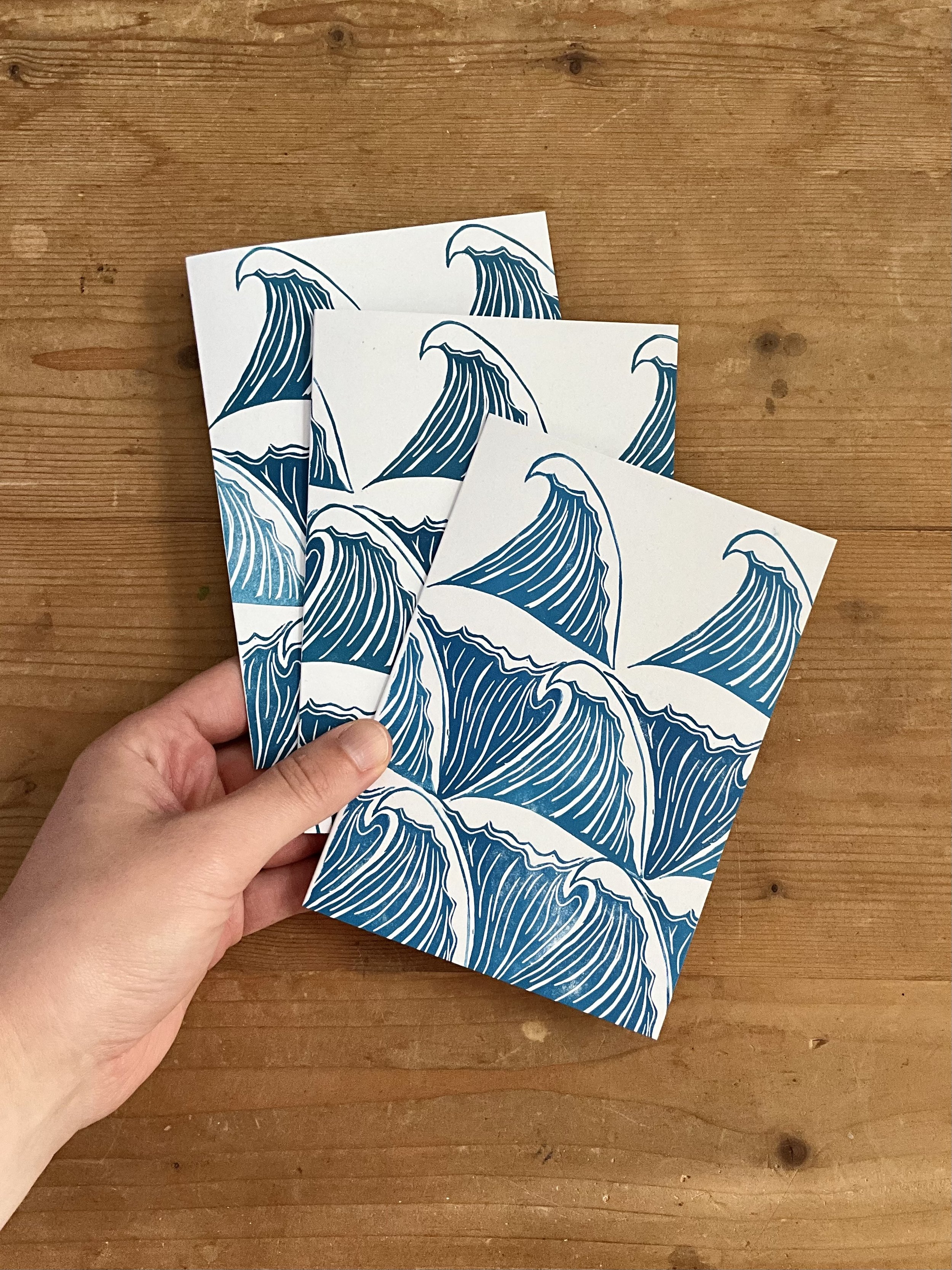 Hand holding three A6 waves linocut greeting cards. These cards have a tessellating waves pattern, printed in teals