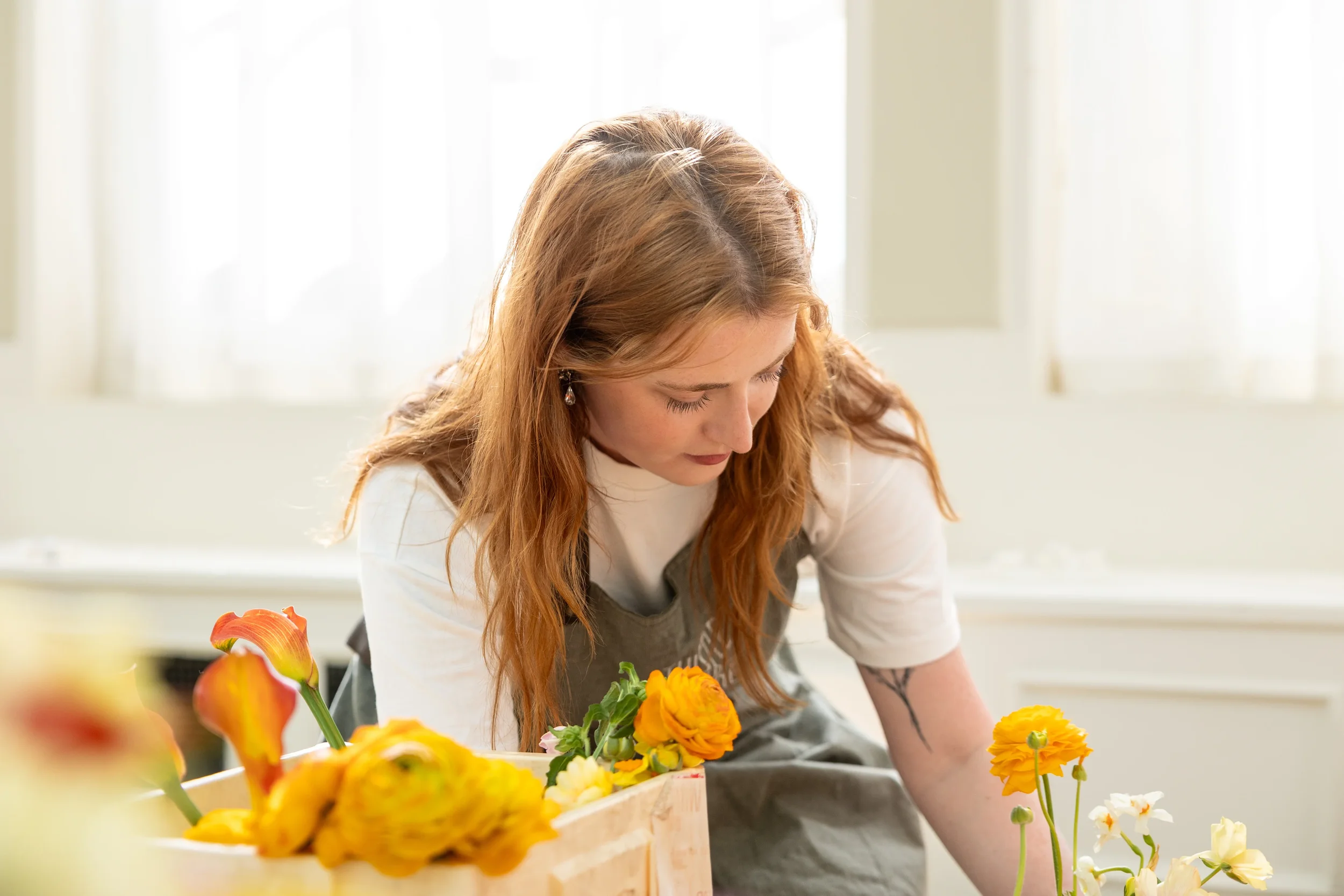 A woman with long red hair working with flowers at a table in a bright room.