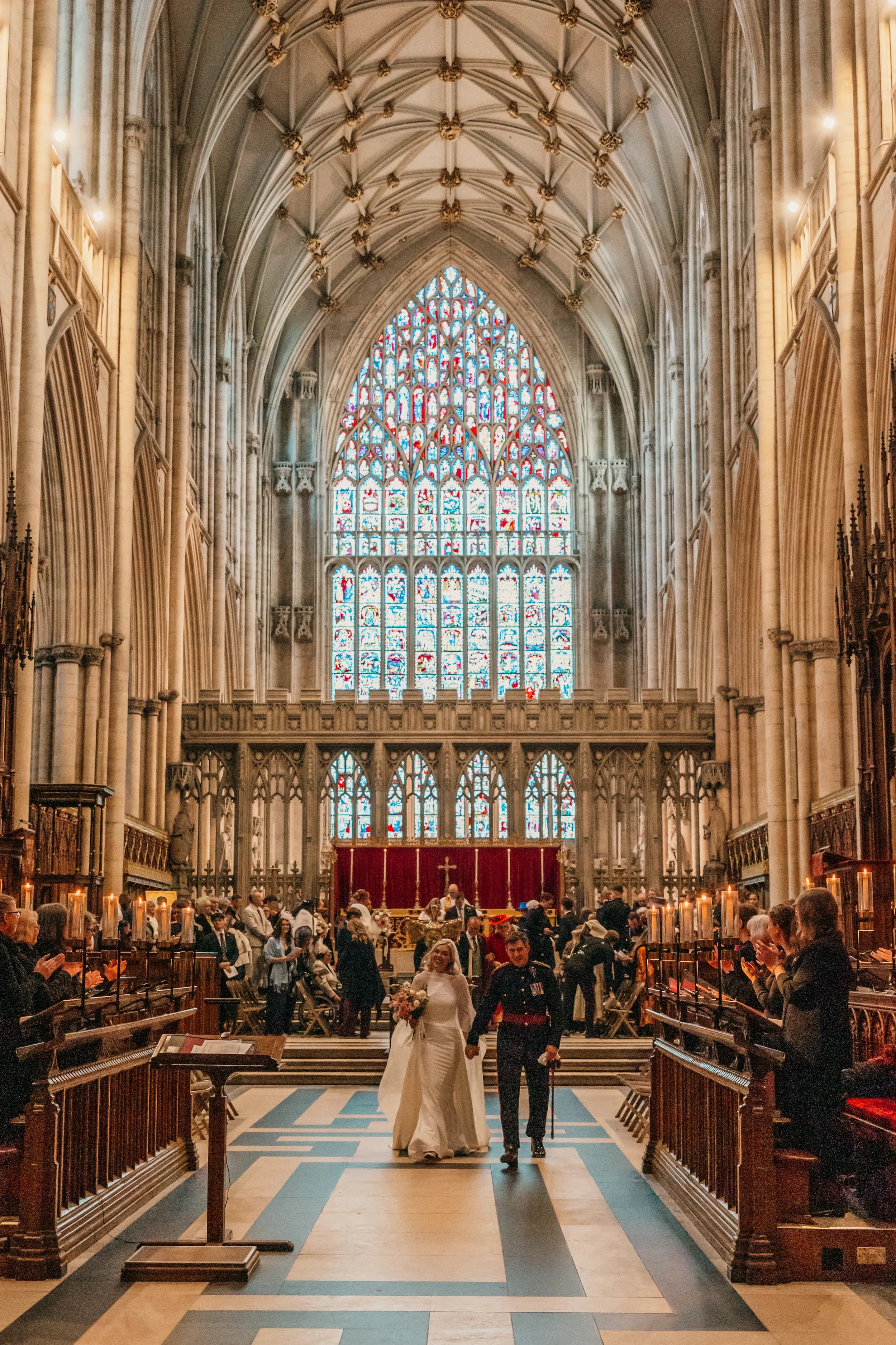 A wedding ceremony inside a large, ornate church with high vaulted ceilings, stained glass windows, and wooden pews. The bride and groom walk down the aisle, surrounded by guests. Candles are lit along the aisle, and the church is decorated for the w