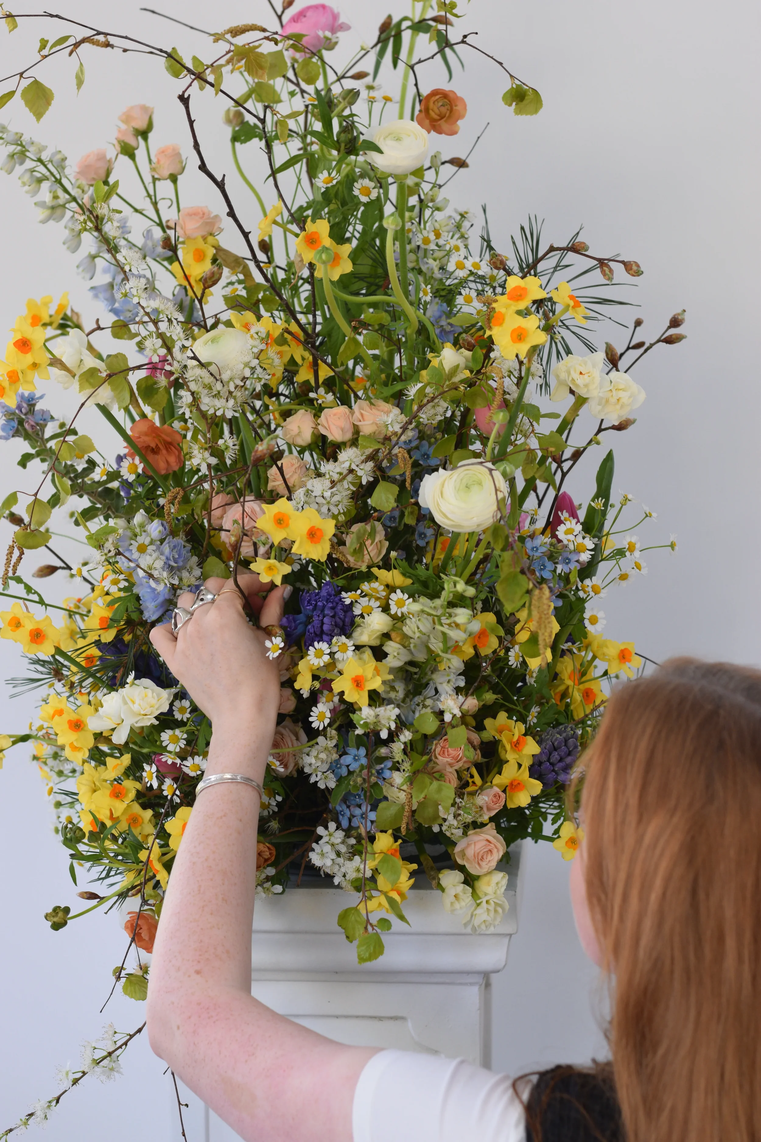 A person with red hair arranging a large and colorful floral arrangement in a white vase. The bouquet contains various flowers including ranunculus, daisies, and other mixed blooms, with a plain white wall as the background.