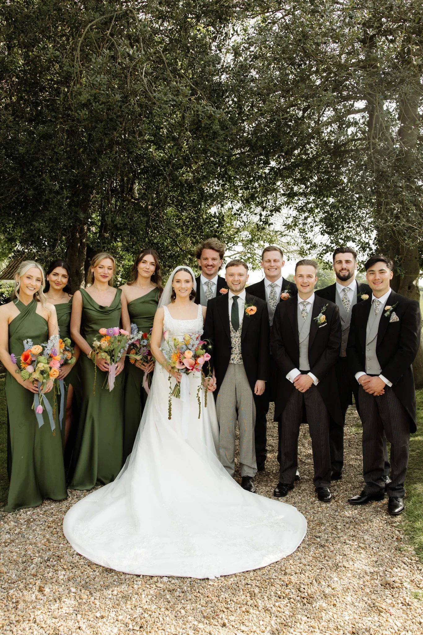 A wedding party standing outdoors with a large tree overhead. The bride and groom are in the center, with bridesmaids in matching green dresses on the left and groomsmen in black suits on the right. They are all smiling and holding colorful bouquets.