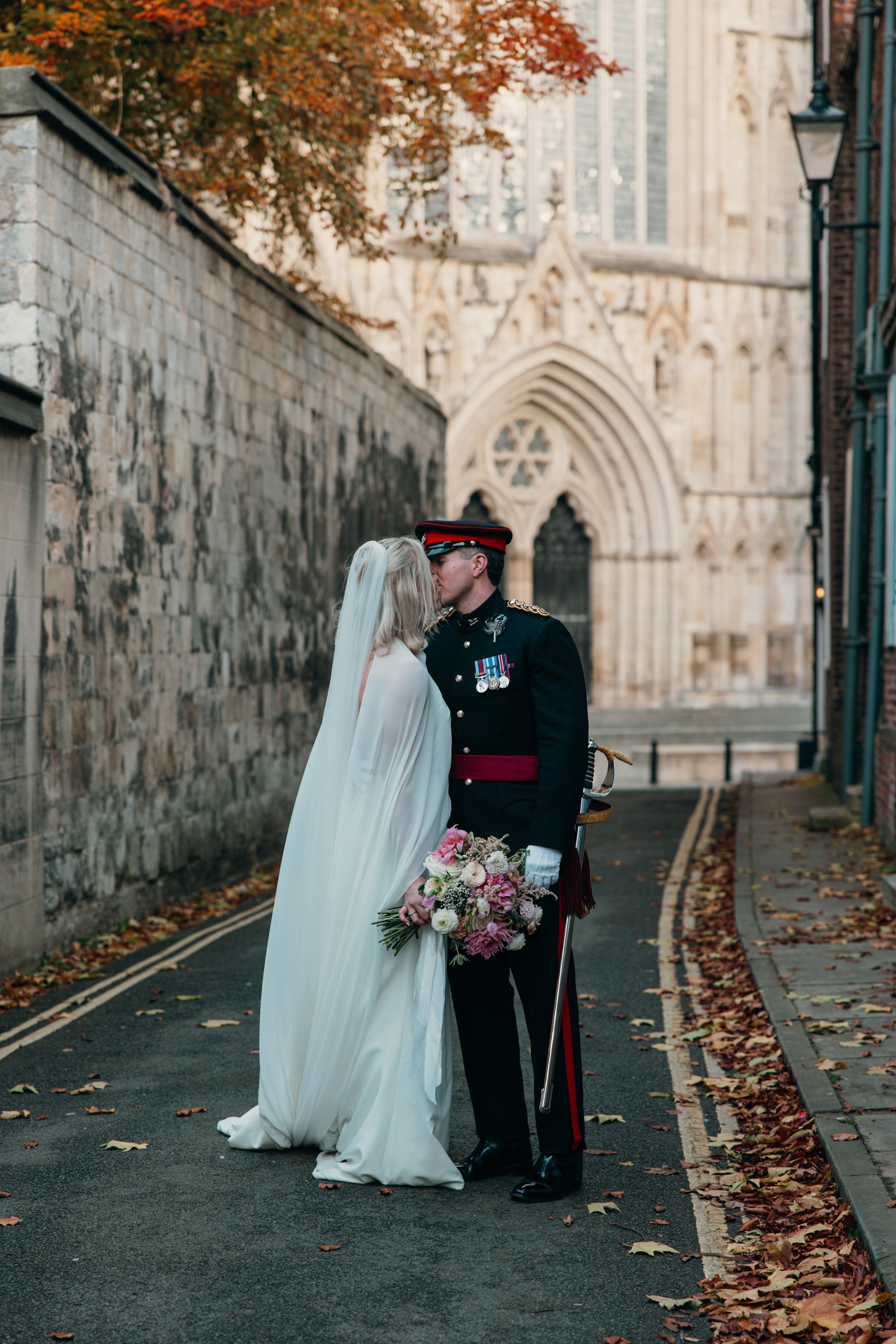 A bride and groom sharing a kiss on a city street with historic architecture in the background, fallen autumn leaves on the road.