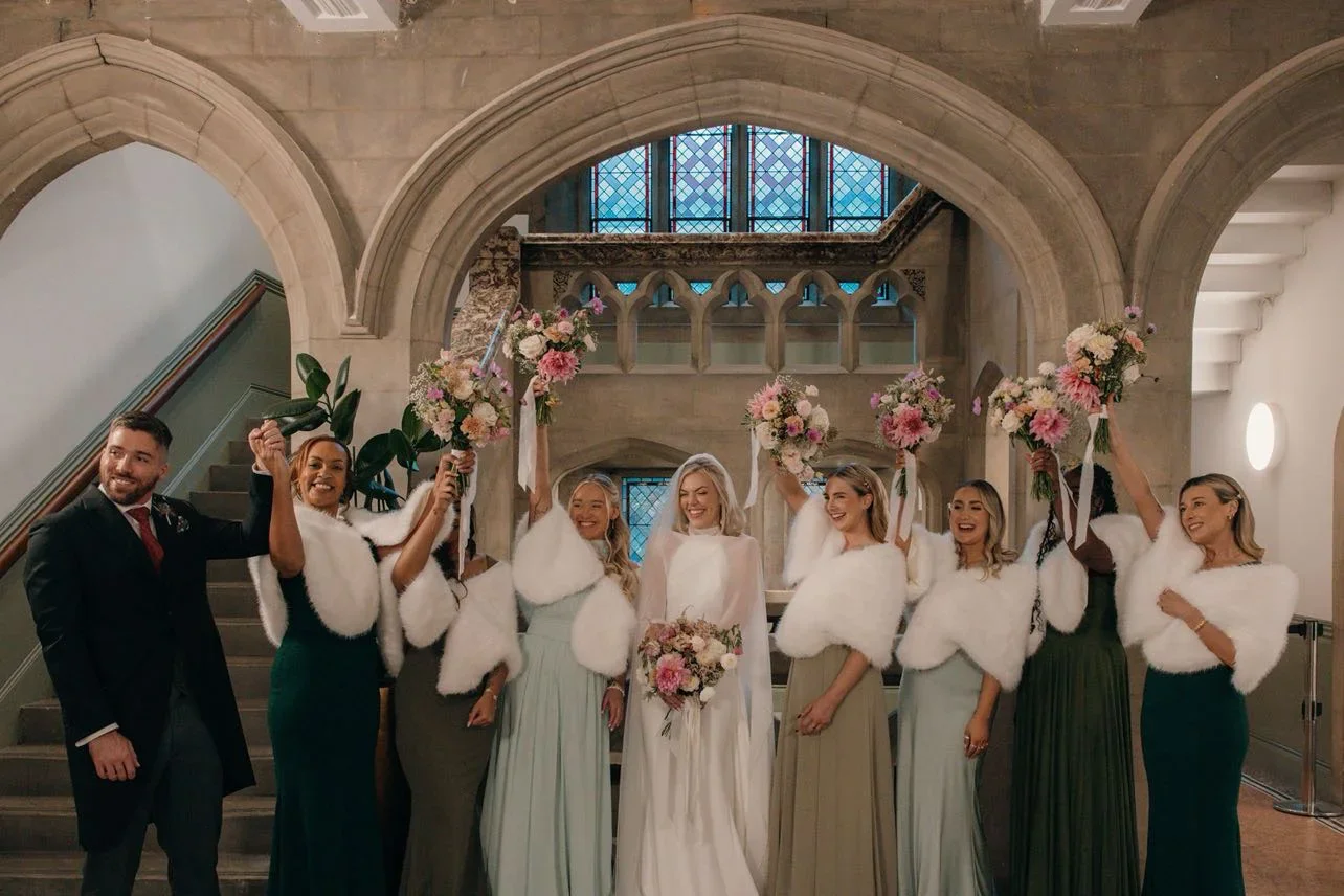 A bride and six bridesmaids, dressed in elegant gowns and white shawls, celebrating in a church with arched architecture and stained glass windows behind them. The bridesmaids are holding pink and white bouquets raised in the air.