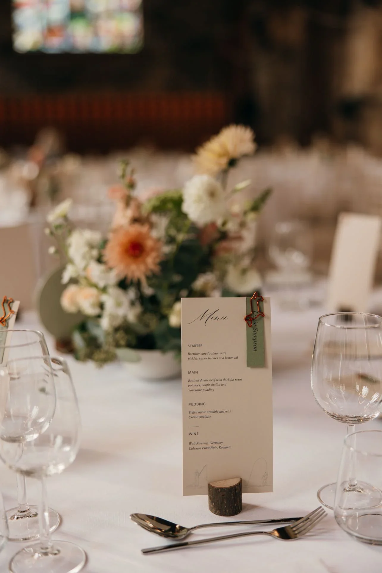 A wedding or formal dinner table setting with a flower centerpiece, a menu card held by a small log piece, and empty wine glasses, with blurred background and a tablet screen displaying colorful images.