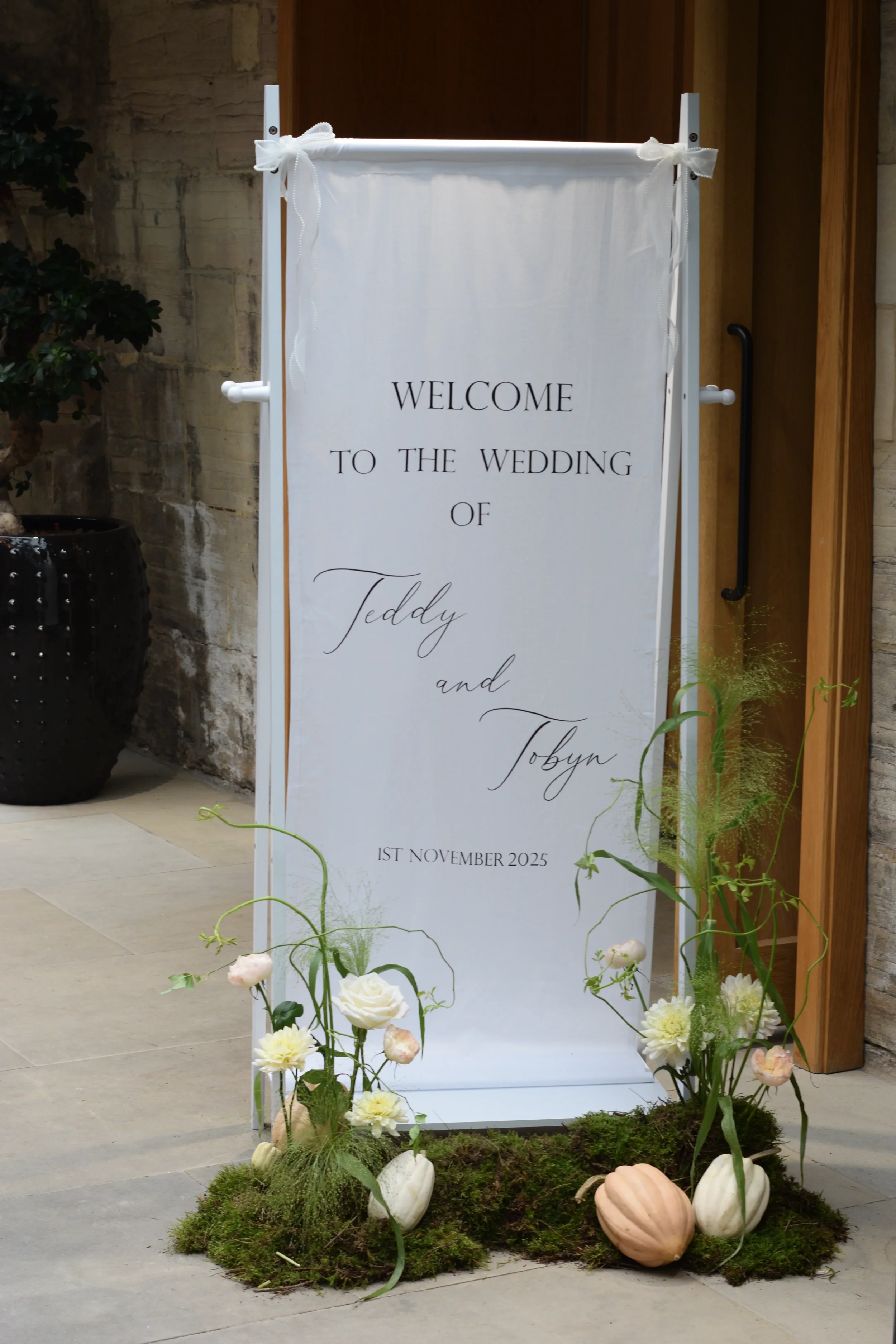 A decorative wedding sign with floral arrangements and pumpkins, welcoming guests to the wedding of Teddy and Tobyn on November 1st, 2025.