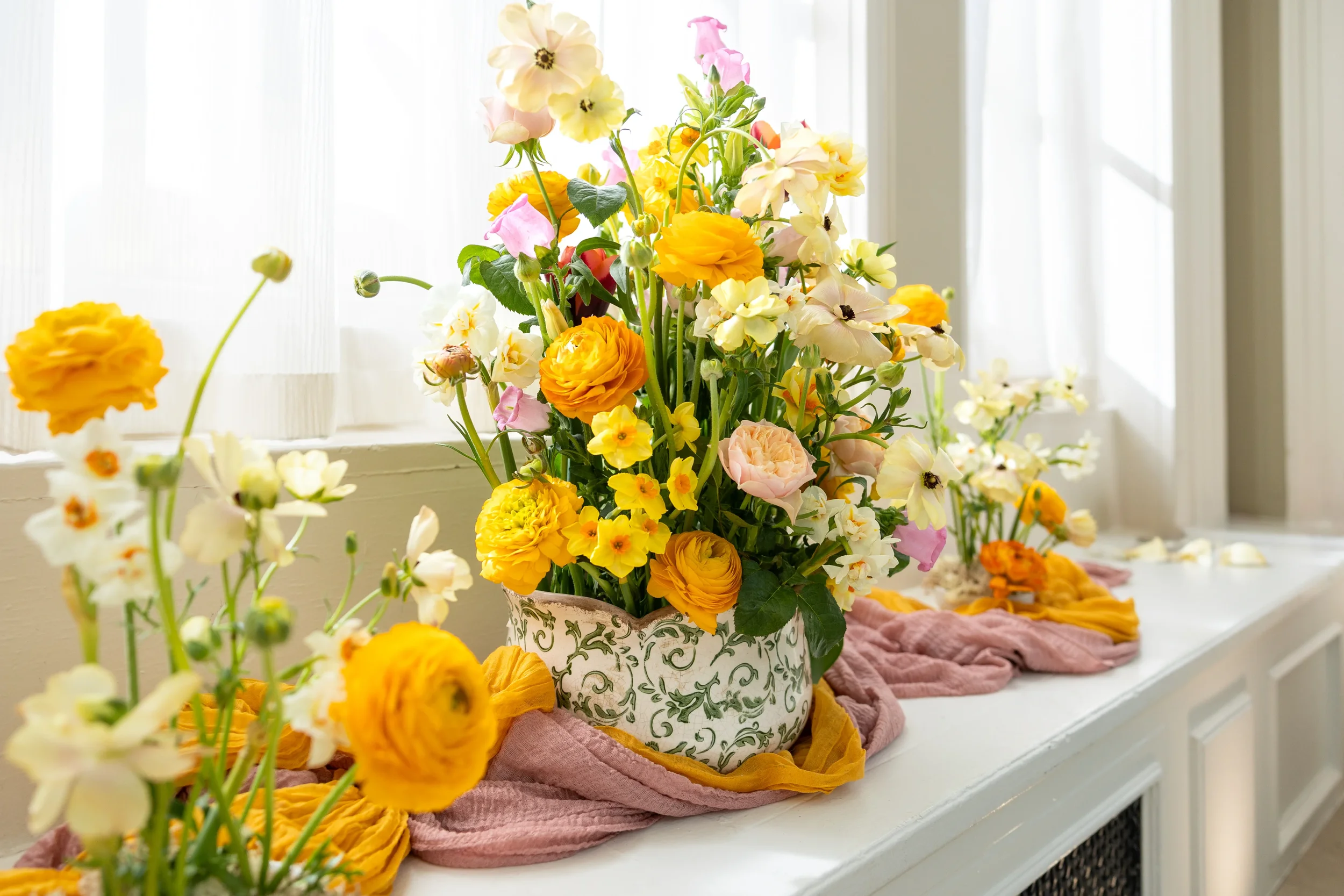 A variety of colorful flowers arranged in a decorative vase on a table, with additional flowers and linens nearby, in a bright room with sheer curtains.