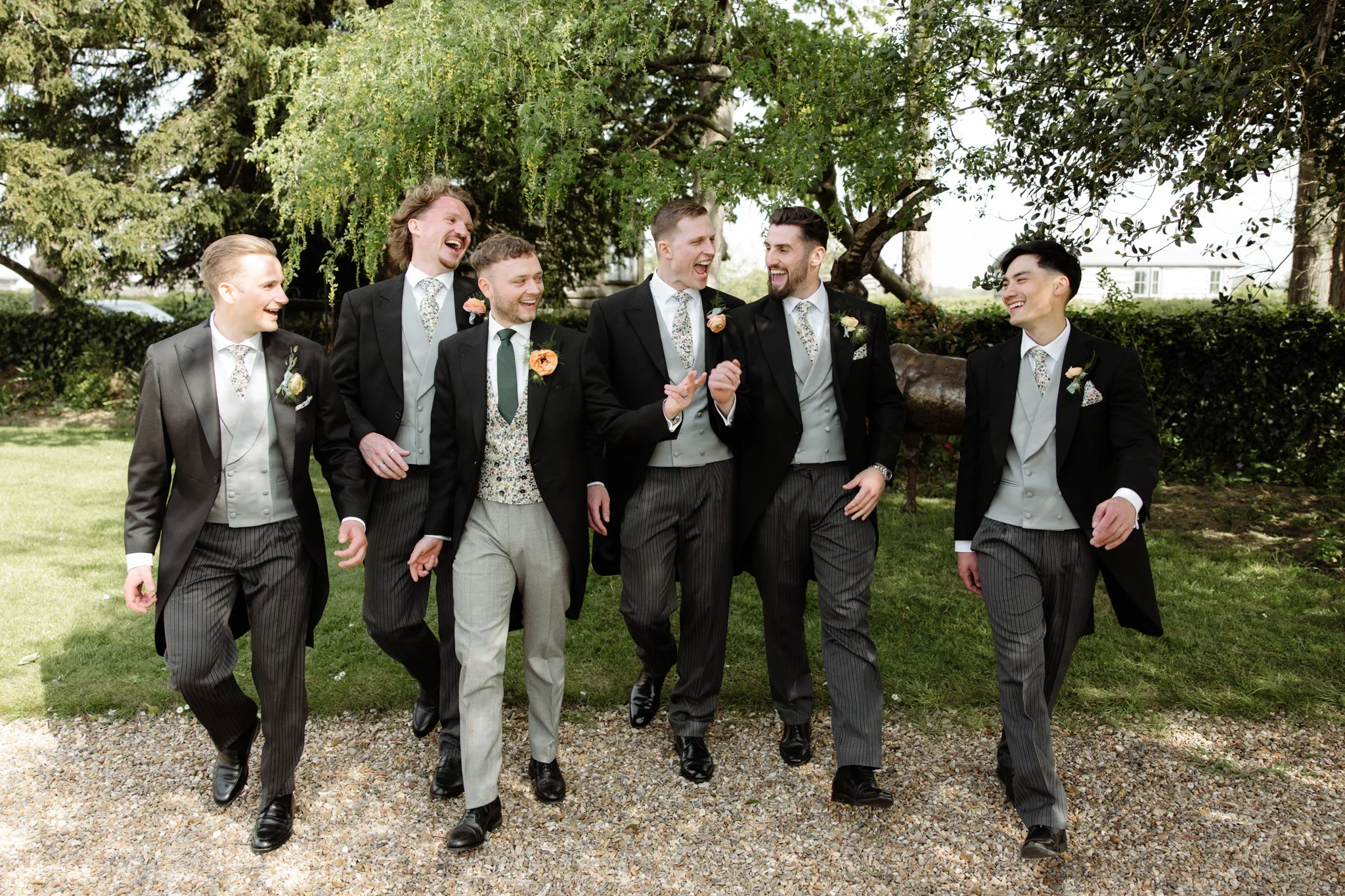 A group of seven men dressed in tuxedos with boutonnières, walking and laughing outdoors in front of trees and greenery, during a wedding celebration.