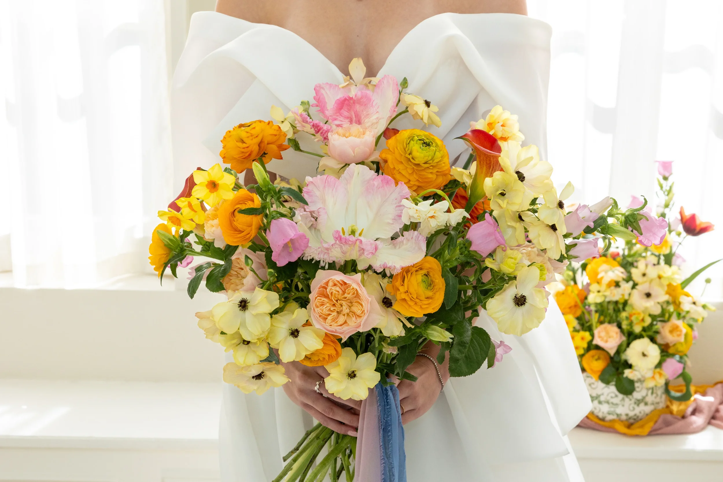 Person holding a large bouquet of mixed pink, yellow, orange, and white flowers, dressed in a white outfit.