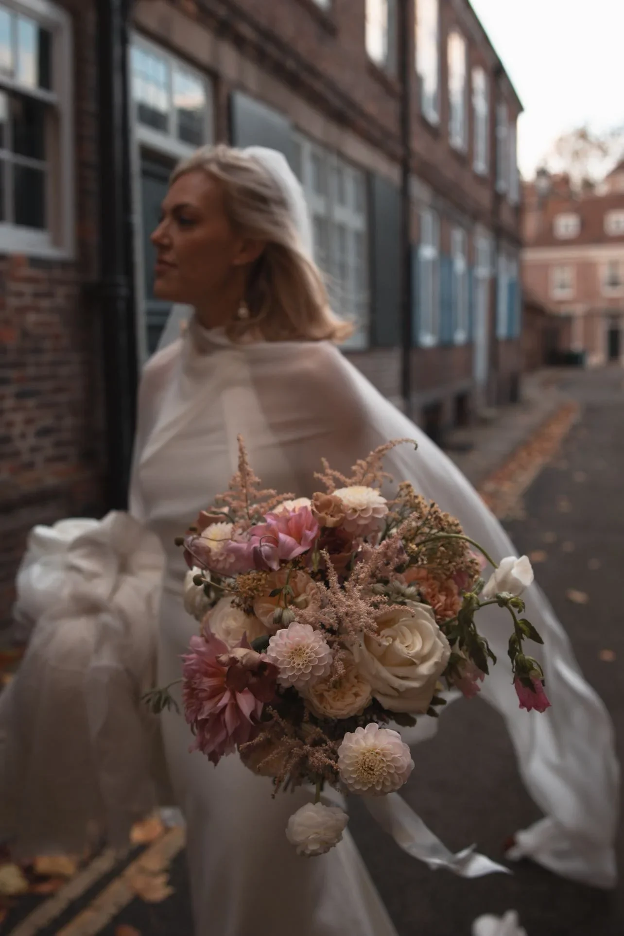 A woman wearing a light-colored dress holding a bouquet of pink and white flowers standing outdoors near a brick building, during late afternoon or early evening.