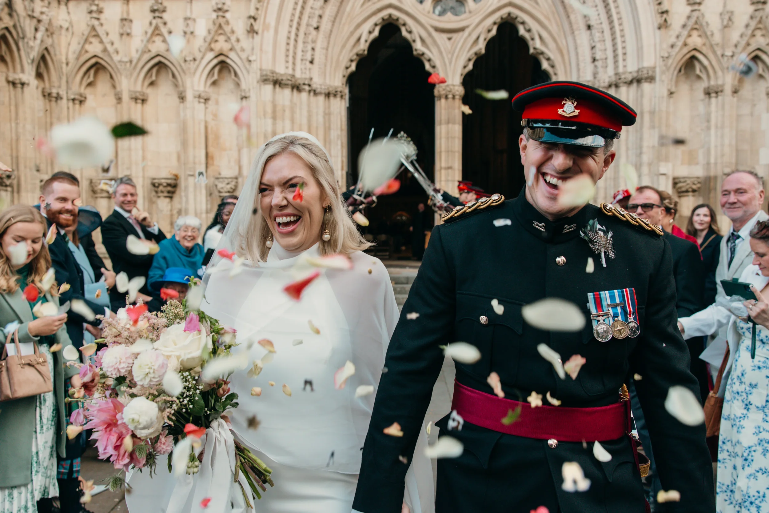Wedding celebration outside a historic church with a bride holding a bouquet of flowers and a man in a military uniform, surrounded by friends throwing flower petals.