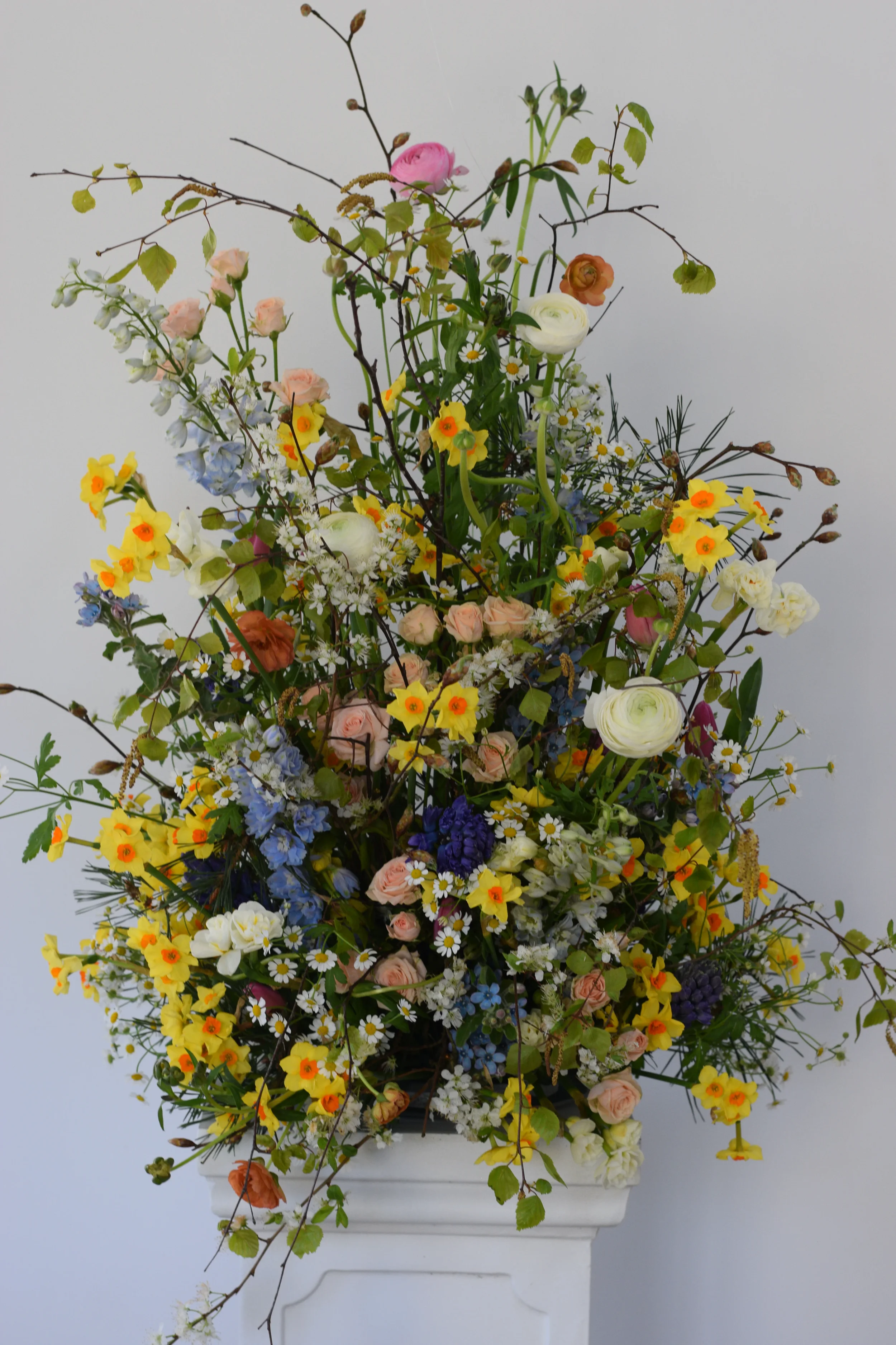A large floral arrangement in a white pedestal vase with mixed flowers including roses, daisies, and wildflowers, against a plain white background.