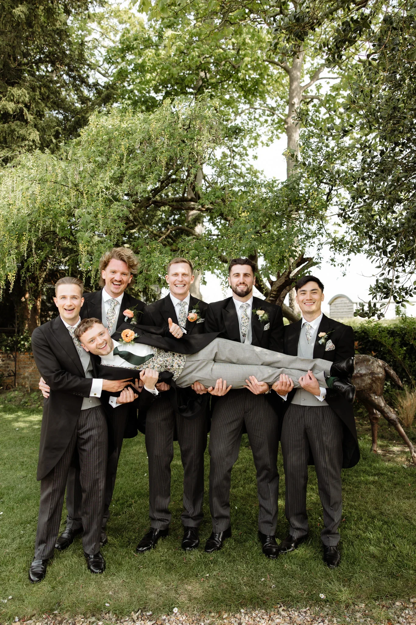 Group of men in tuxedos lifting another man in a wedding pose outdoors with trees in the background.
